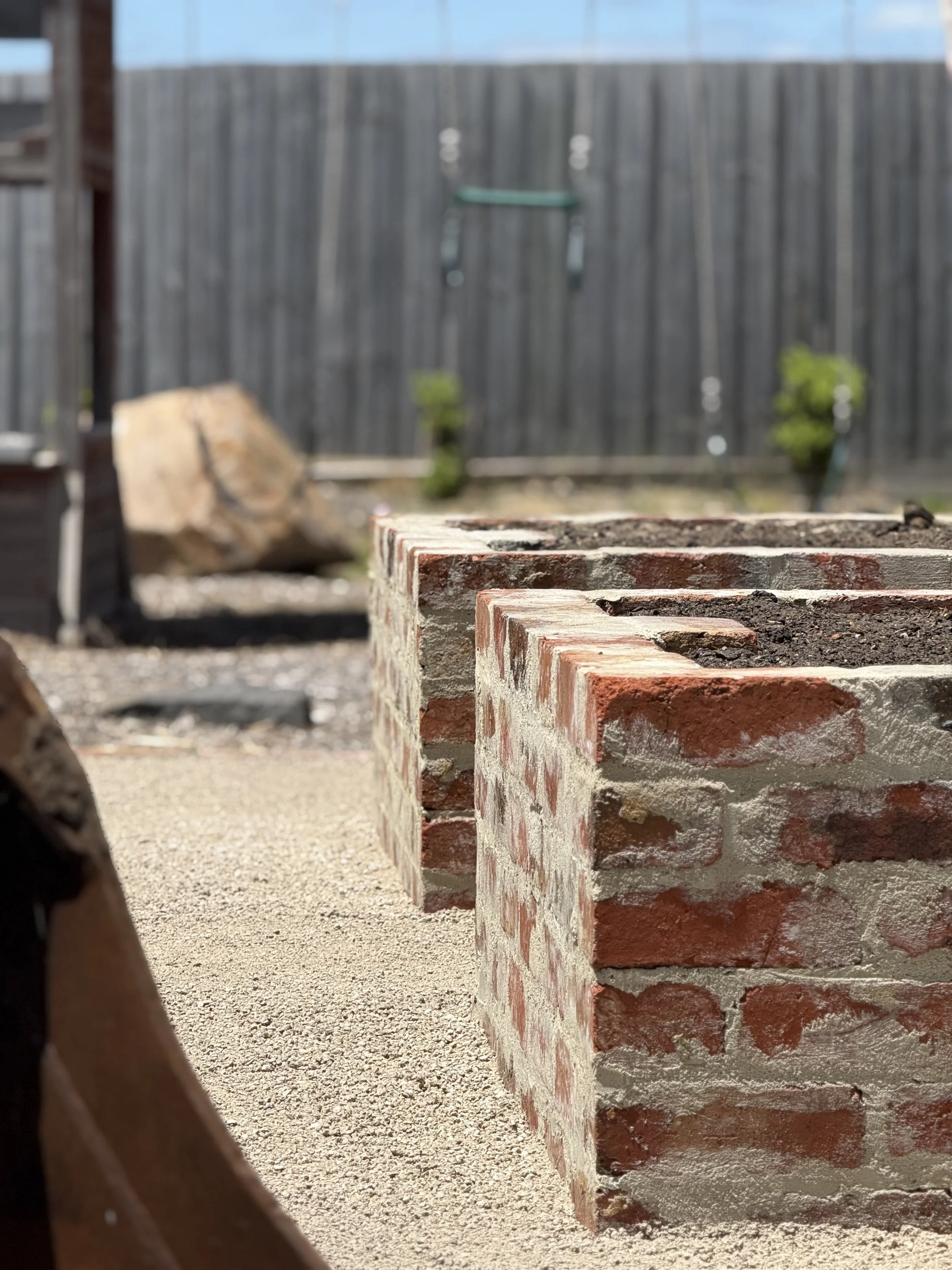 Close-up view of red brick planters with soil, in a backyard with a wooden fence and small plants in the background.
