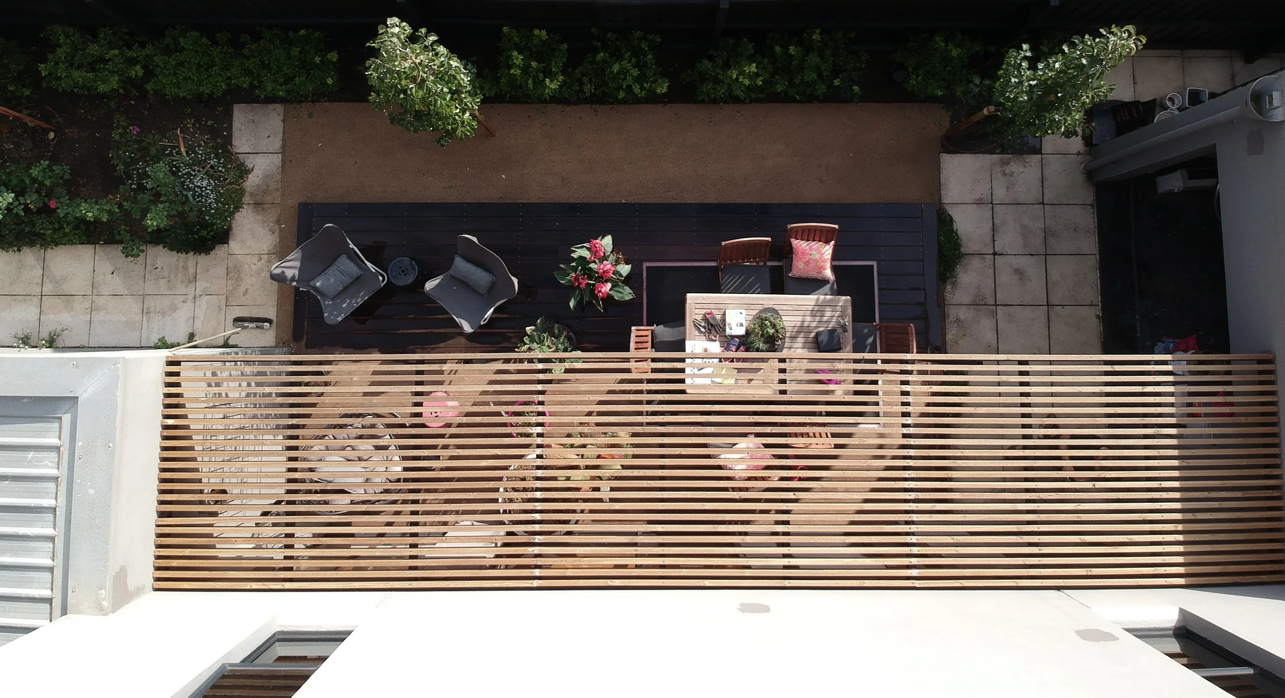 Top-down view of a balcony with outdoor furniture, potted plants, a table with items, sunlit wooden slats, and a paved and garden area with trees and bushes.