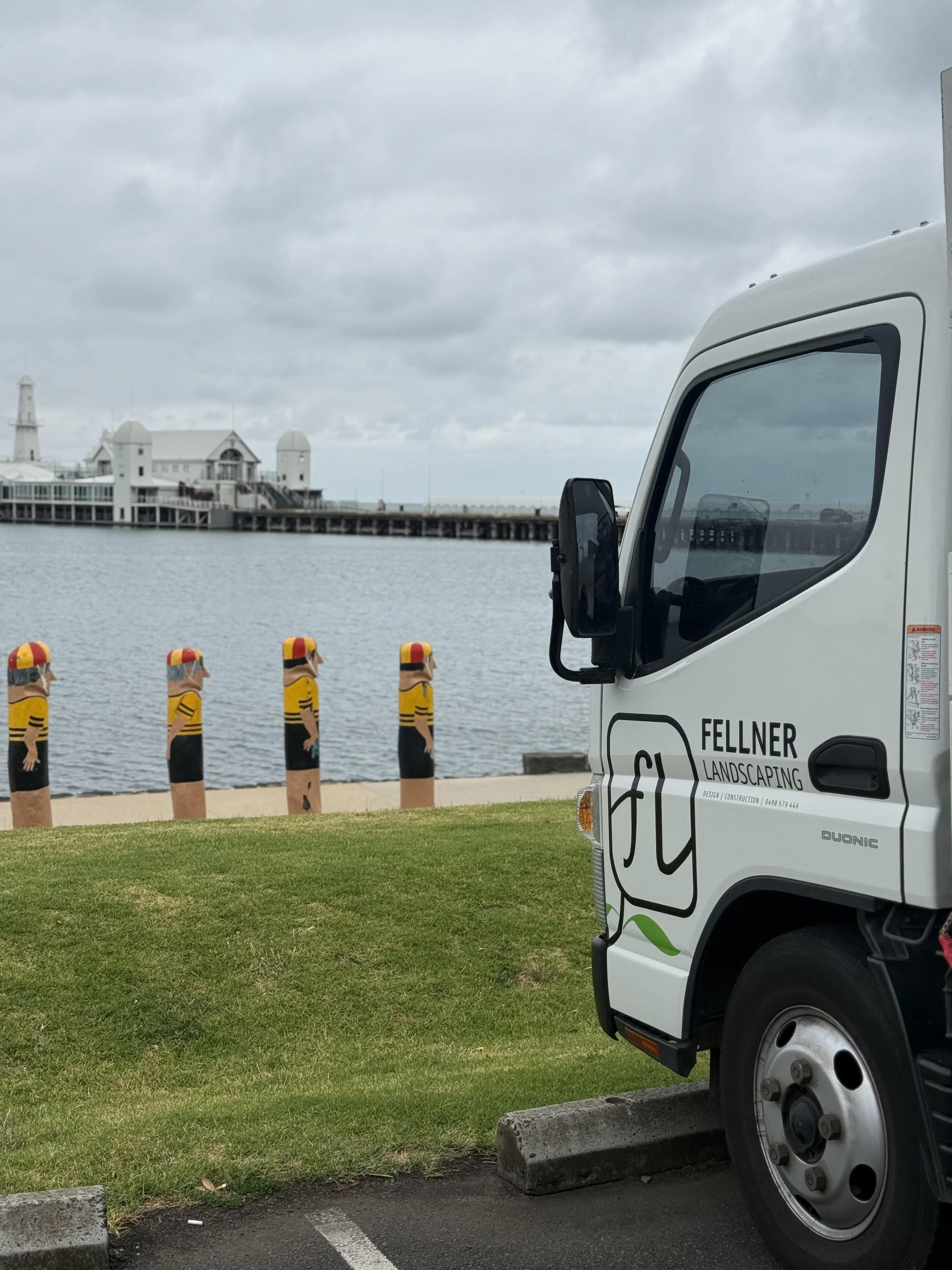 A white landscaping truck parked near a grassy area by the water, with four yellow and black painted bollards featuring cartoonish figures in helmets, a pier, and a building with a lighthouse in the background under cloudy skies.