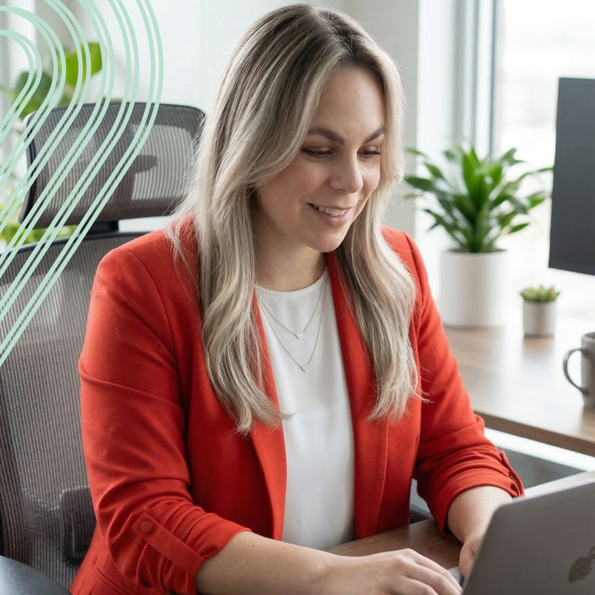 A woman with long blonde hair wearing a red blazer and white top, sitting at a desk, looking at a laptop, in an office with plants and large windows.