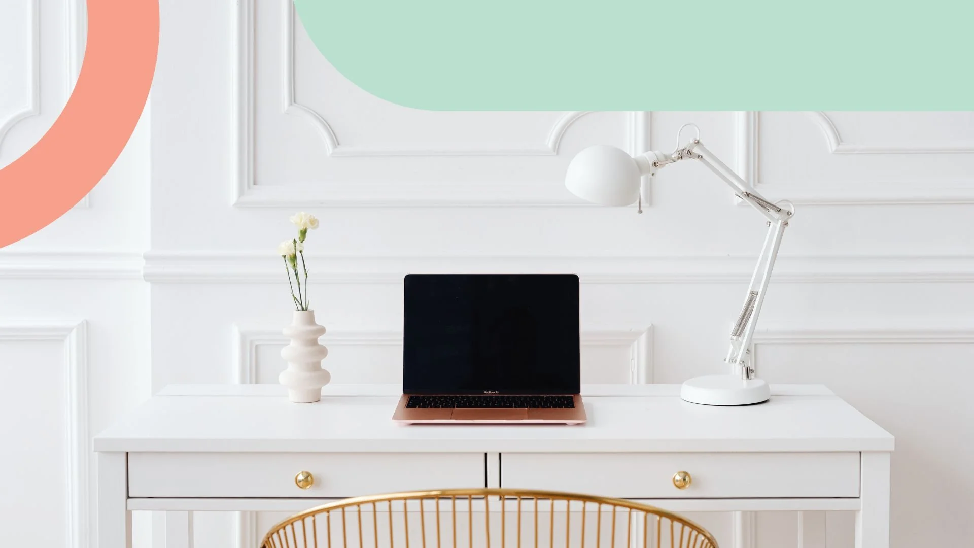 White desk with a black laptop, white desk lamp, and a white vase with yellow and white flowers against a paneled white wall.