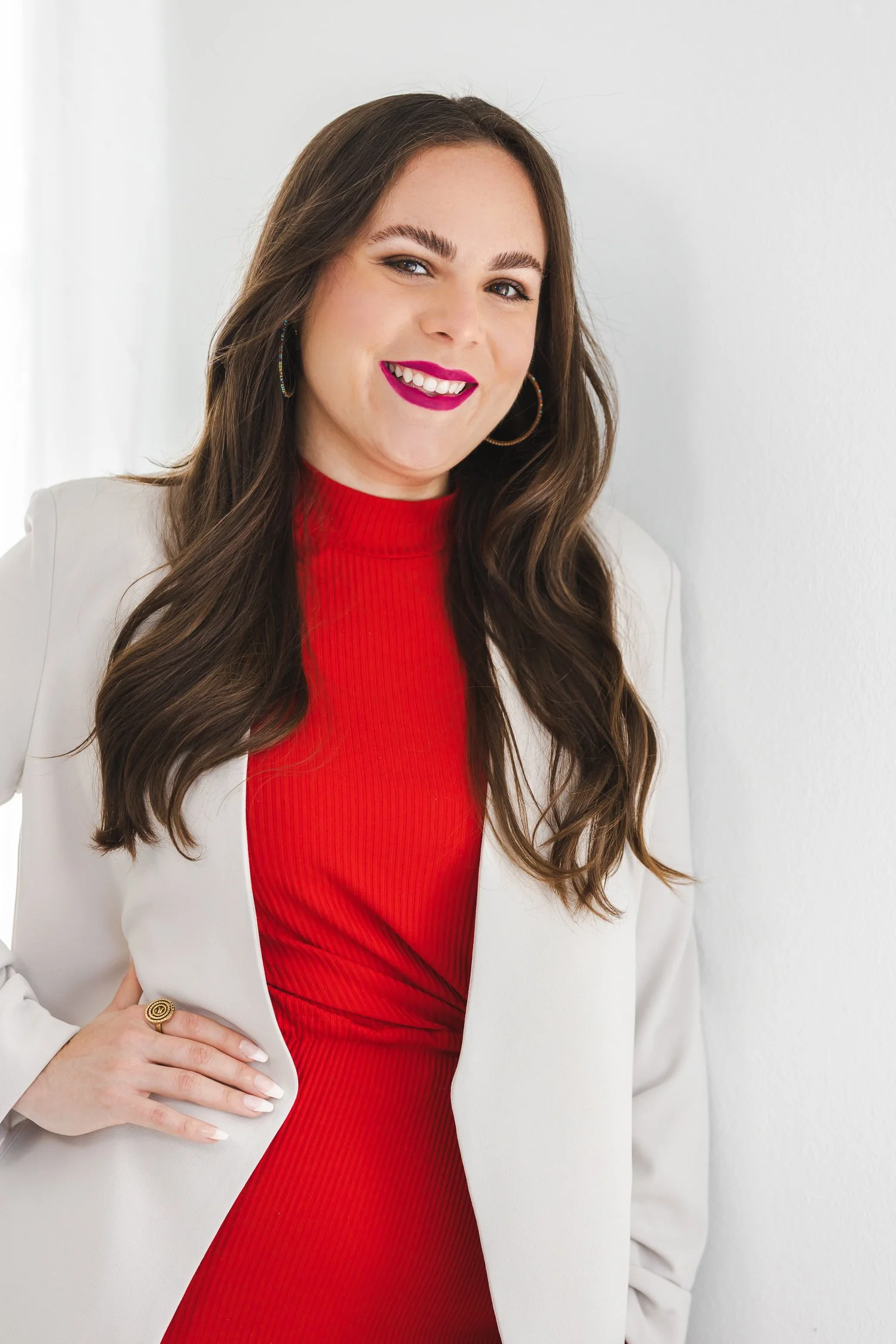 A woman with long brown hair wearing a red dress, white blazer, hoop earrings, and pink lipstick, smiling and standing against a white wall.
