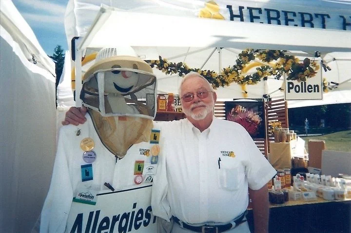 A man and a beekeeper statue posing together at an outdoor event booth. The man has white hair and a beard, wearing glasses and a white shirt. The booth has a sign with the word 'Pollen' and various honey products displayed.
