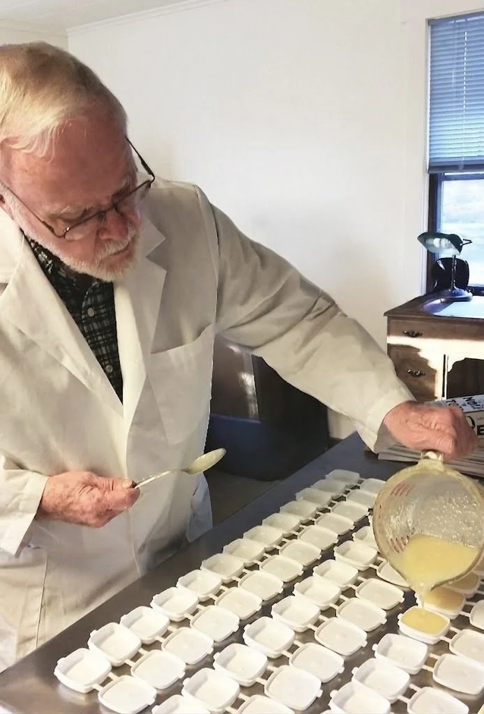 An elderly man in a white coat is pouring yellow liquid into small, white, square molds placed on a table, with many already filled molds.