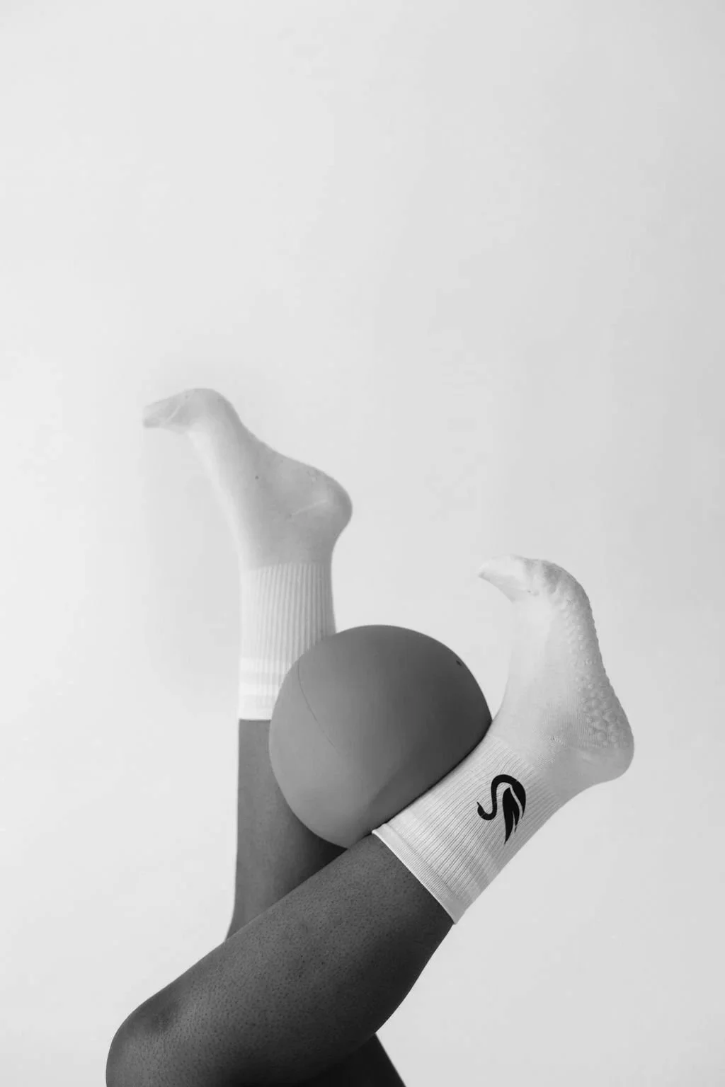 A black and white photo showing a person’s leg wearing a sock with a logo, holding a ping pong ball between their foot and a hand, against a plain background.