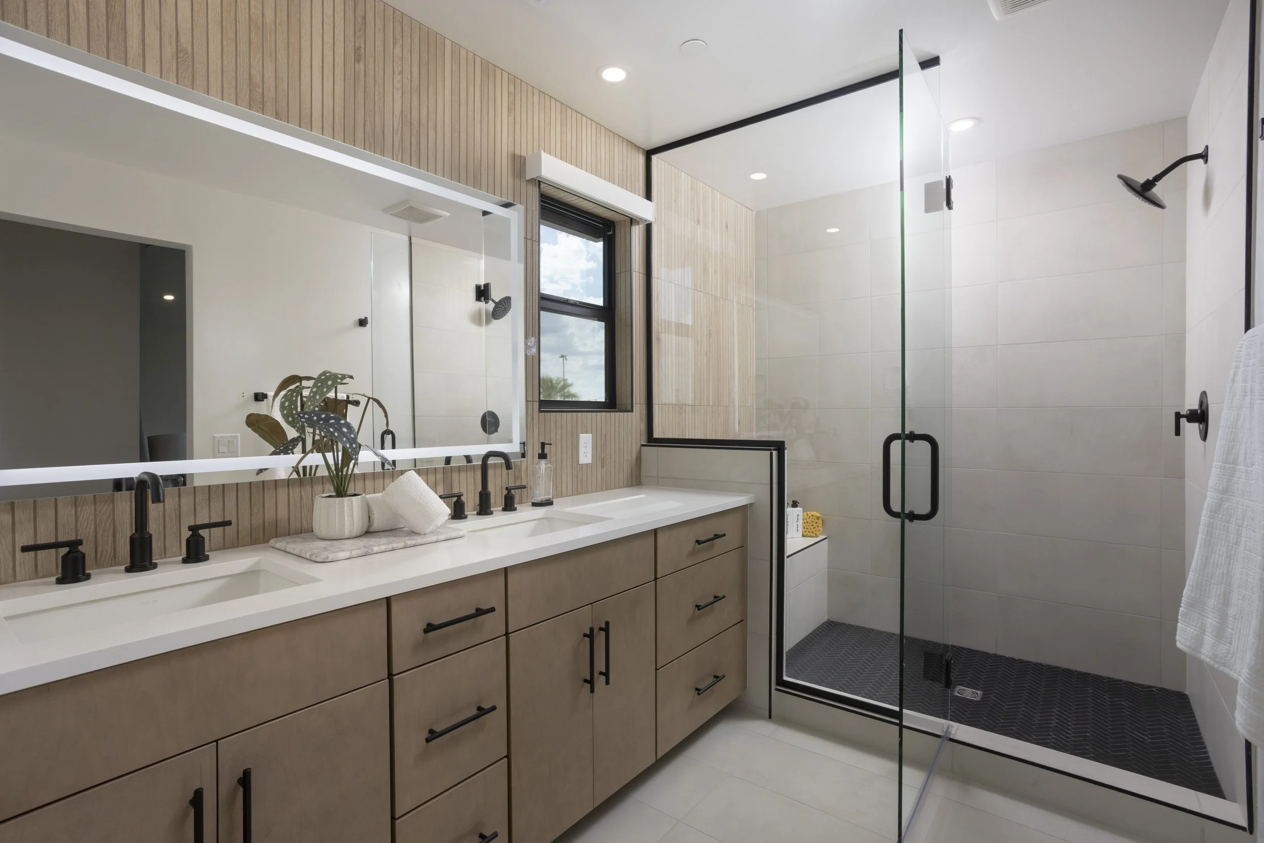 Modern bathroom featuring a double sink vanity with wooden drawers, black faucets, decorative plants, large mirror, glass-enclosed shower with black fixtures, white tiled walls, and a window with outdoor view.