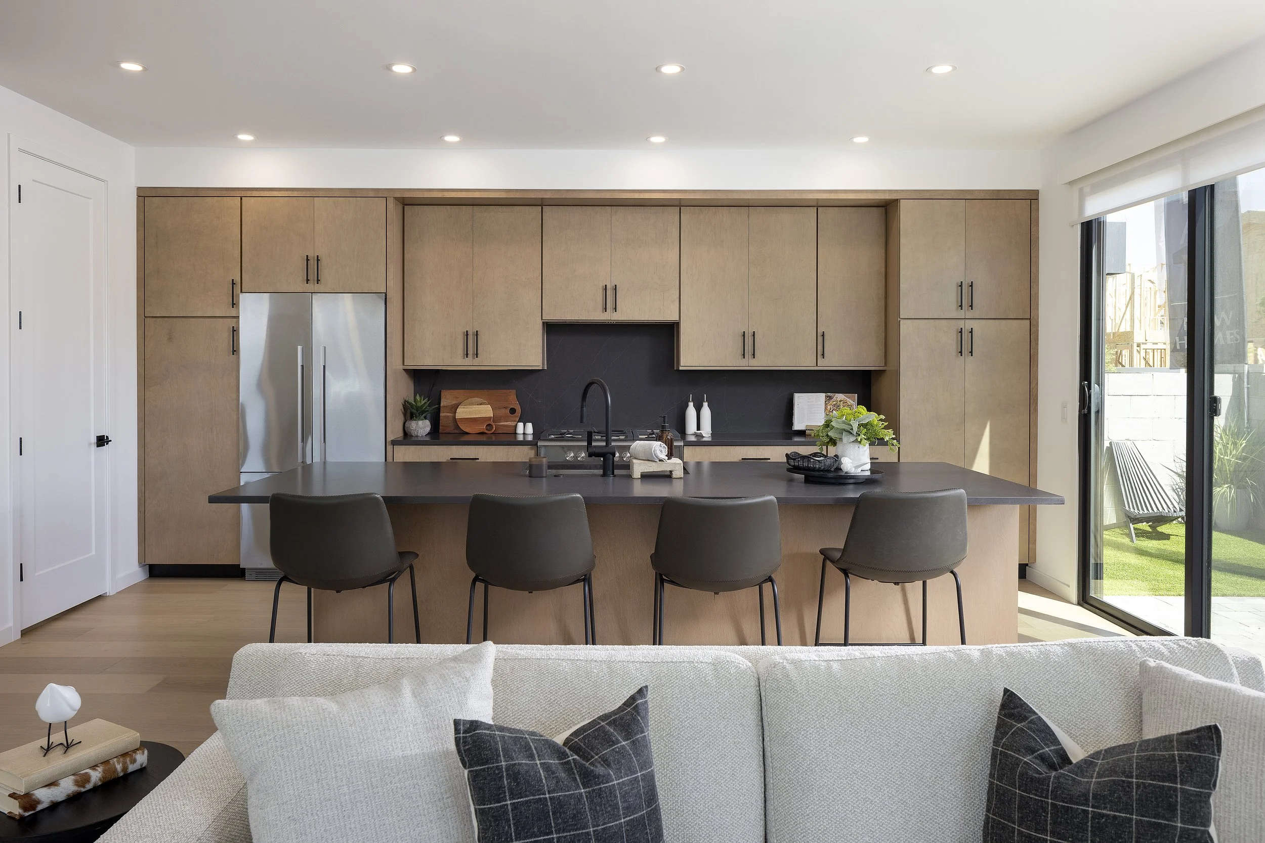 Modern kitchen with light wood cabinets, black countertop, stainless steel refrigerator, black sink, and bar stools facing a living room with a beige sofa and checkered pillows.