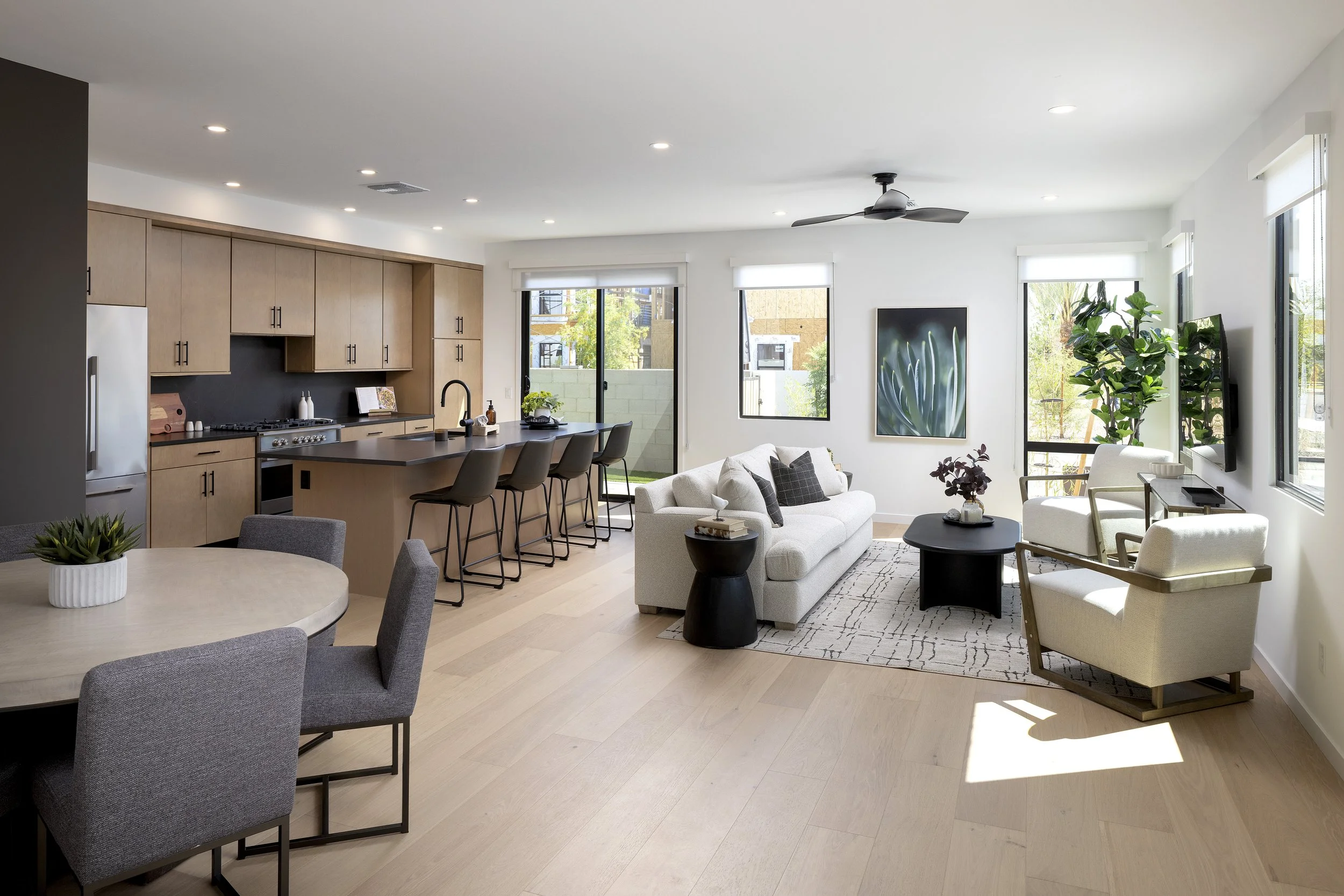 Open-concept living room and kitchen with light wood flooring, white walls, large windows, ceiling fan, beige sofa, armchair, black coffee table, potted plants, and a kitchen island with bar stools.