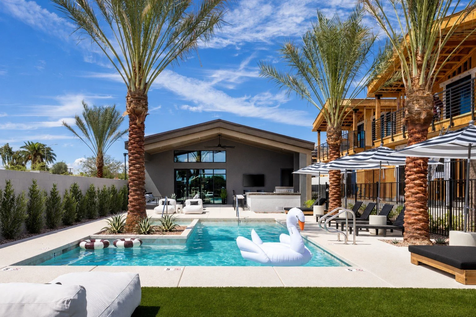 A modern luxury complex outdoor swimming pool area with lounge chairs, umbrellas, and a fire feature, surrounded by landscaped plants and trees.