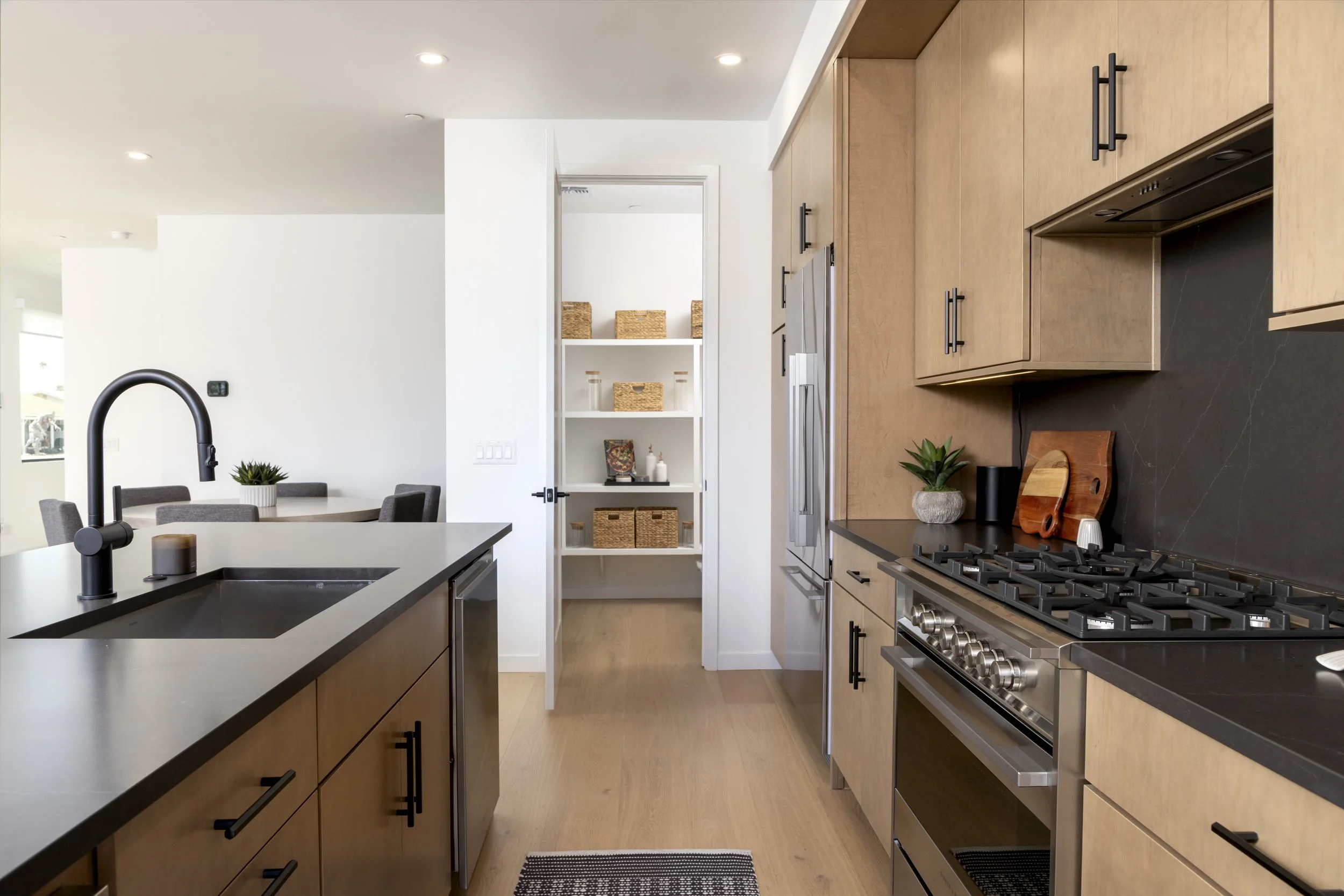 Modern kitchen with black countertops, wooden cabinets, stainless steel appliances, and a black stove, with a dining area visible in the background.
