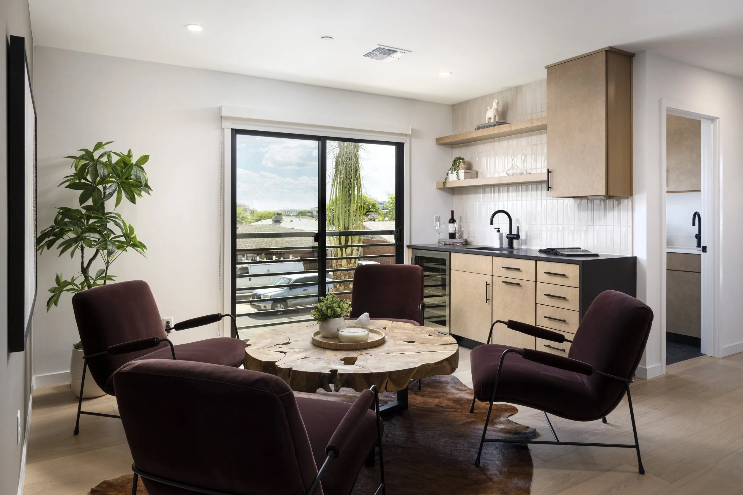 A living room with a round wooden coffee table surrounded by five maroon velvet armchairs, a Juliet Balcony with sliding glass door, a potted plant, and a kitchen area with light wood cabinets, open shelves, and a black faucet.