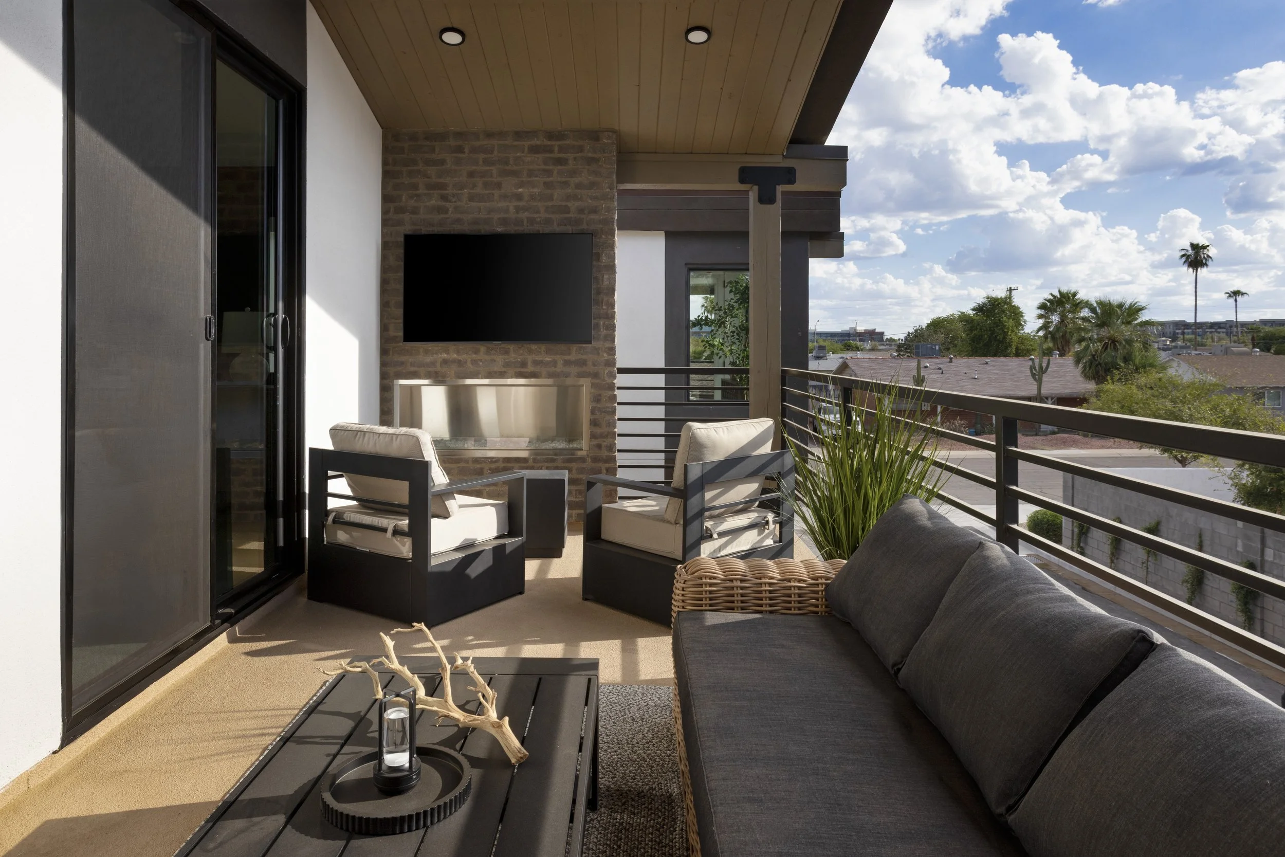 Outdoor balcony with beige carpet, black metal coffee table, black and beige chairs, gray sofa with black cushions, firepit, potted plant, brick fireplace, mounted black flat-screen TV, sliding glass door, wooden ceiling, view of rooftops, palm trees