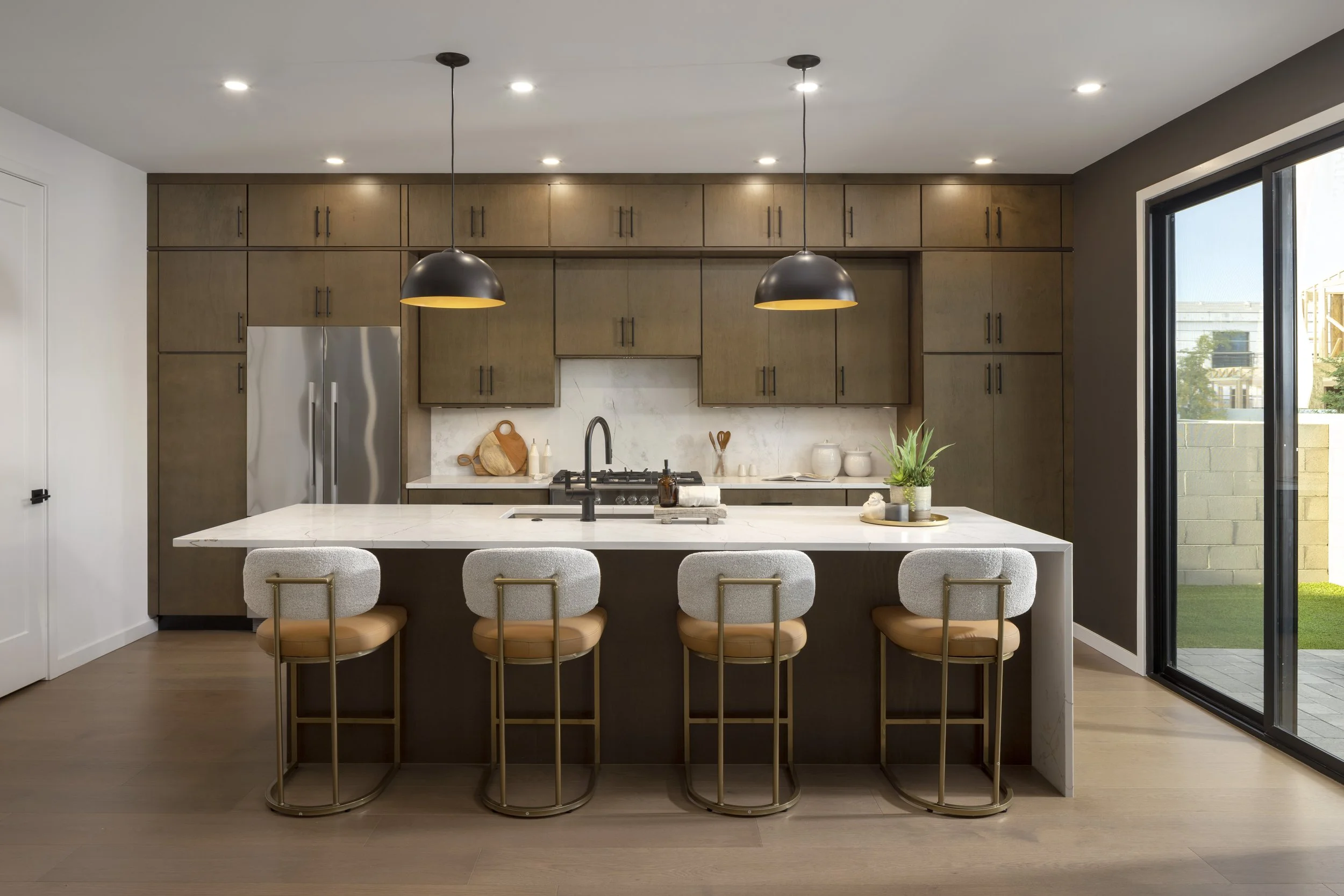 Modern kitchen with dark wood cabinets, stainless steel refrigerator, white marble backsplash, and an island with four gray barstools. Recessed and pendant lighting, light-colored walls, and hardwood floors.