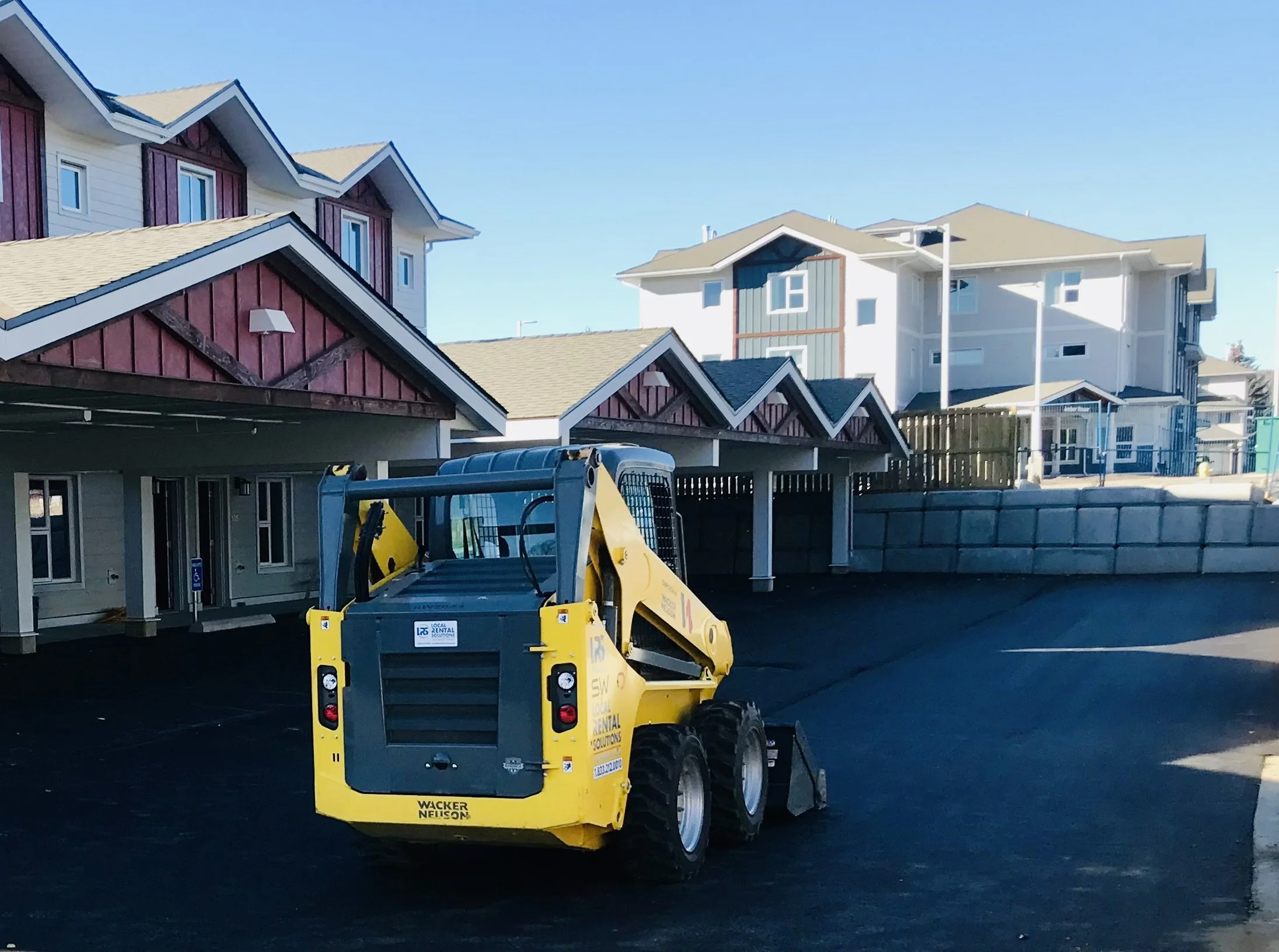 A small construction vehicle parked on freshly laid asphalt in a residential area with multi-story houses in the background.