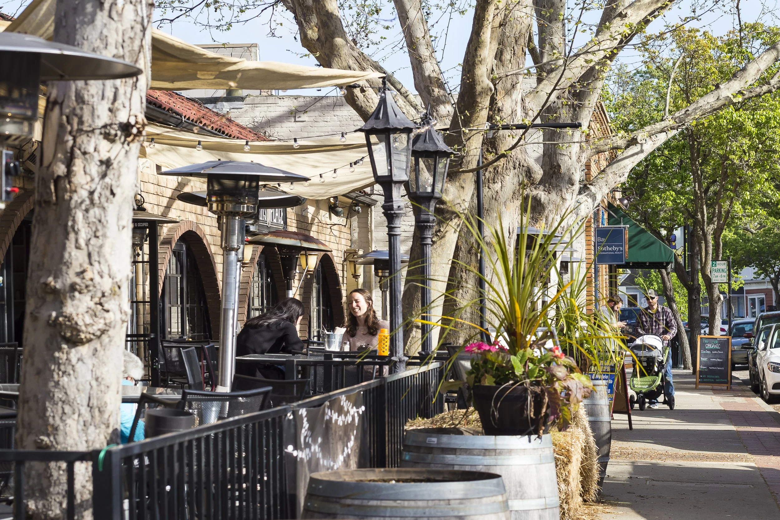 Outdoor patio scene with people sitting at tables, several large trees, string lights, and a street with cars.