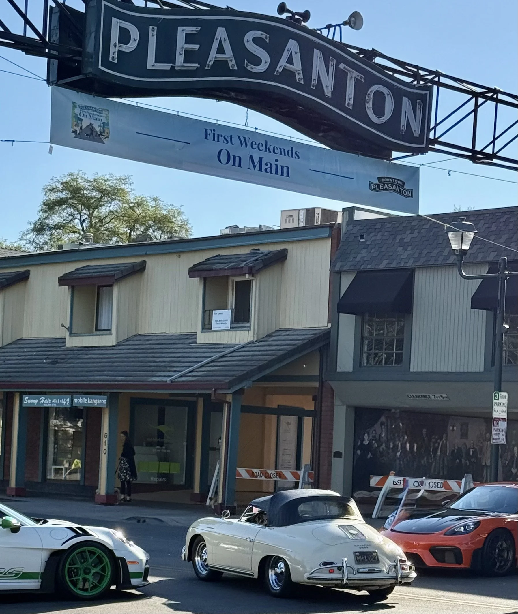 View of a downtown Pleasanton sign with a banner underneath advertising 'First Weekends On Main'. Below, multiple cars are parked, including a white Corvette, a yellow sports car, and an orange sports car, with a mix of buildings and a tree in the ba