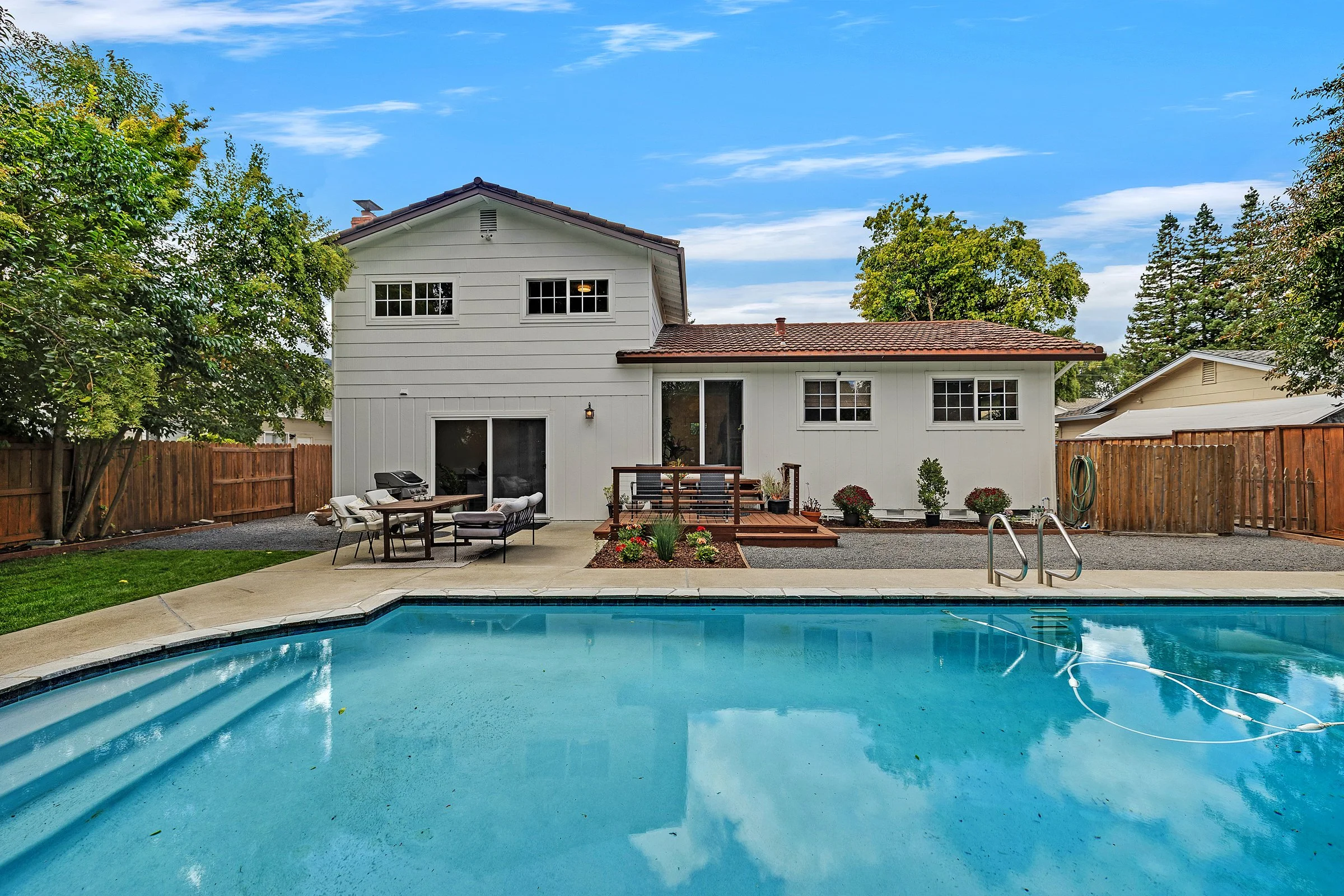 Backyard with swimming pool, patio furniture, and house with deck, surrounded by trees and fenced yard.