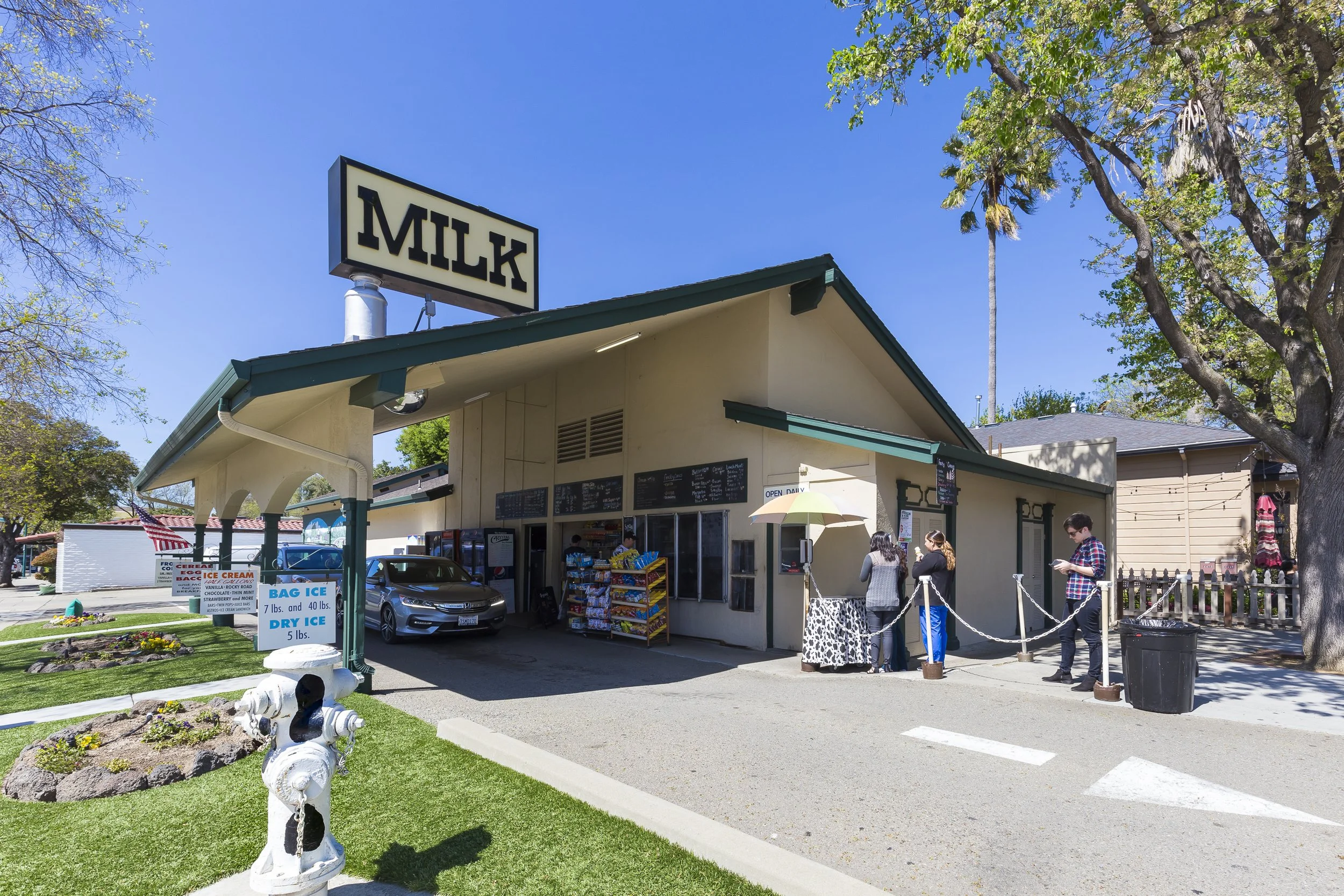 A small cream-colored building with a green roof and a large sign that says 'MILK' on top. In front of the building, there is a parking area with a gray car. Outside, there are snack racks, a sign displaying ice cream and ice prices, and several peop