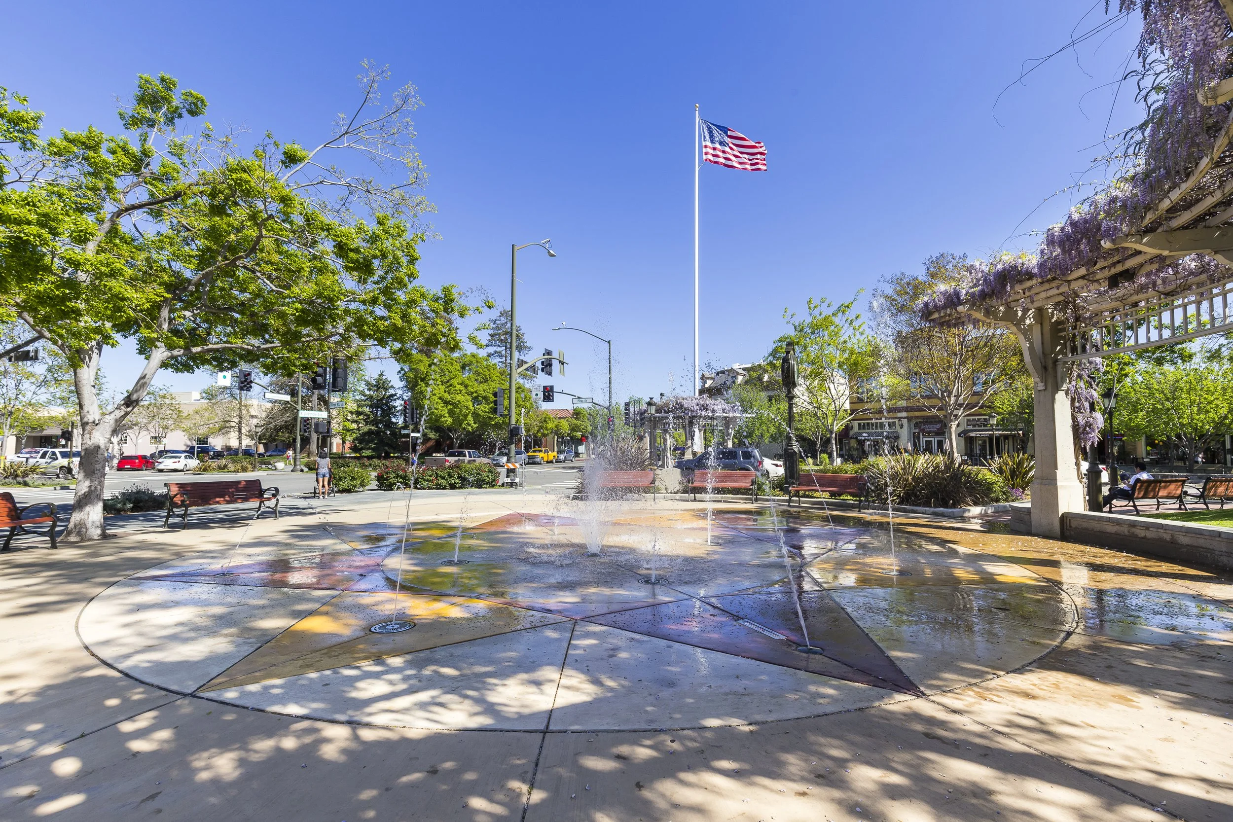 A town square with trees, benches, and a fountain with water spraying from multiple jets. An American flag is flying on a tall flagpole. There are buildings and parked cars in the background, along with traffic lights and street signs.