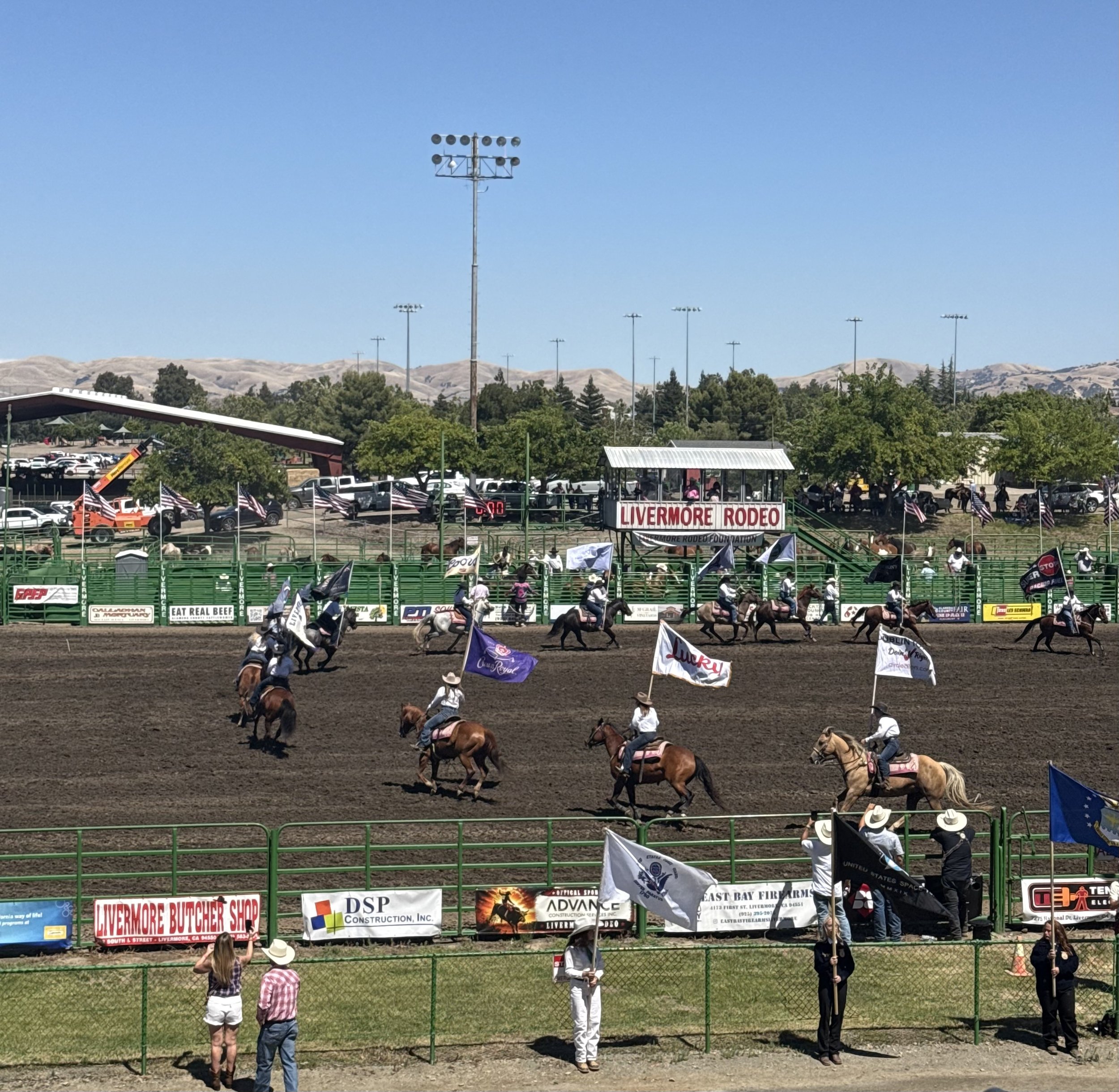 Rodeo event with riders on horseback holding flags, spectators watching from the stands, and banners advertising various local businesses, set against a backdrop of hills and a clear blue sky.