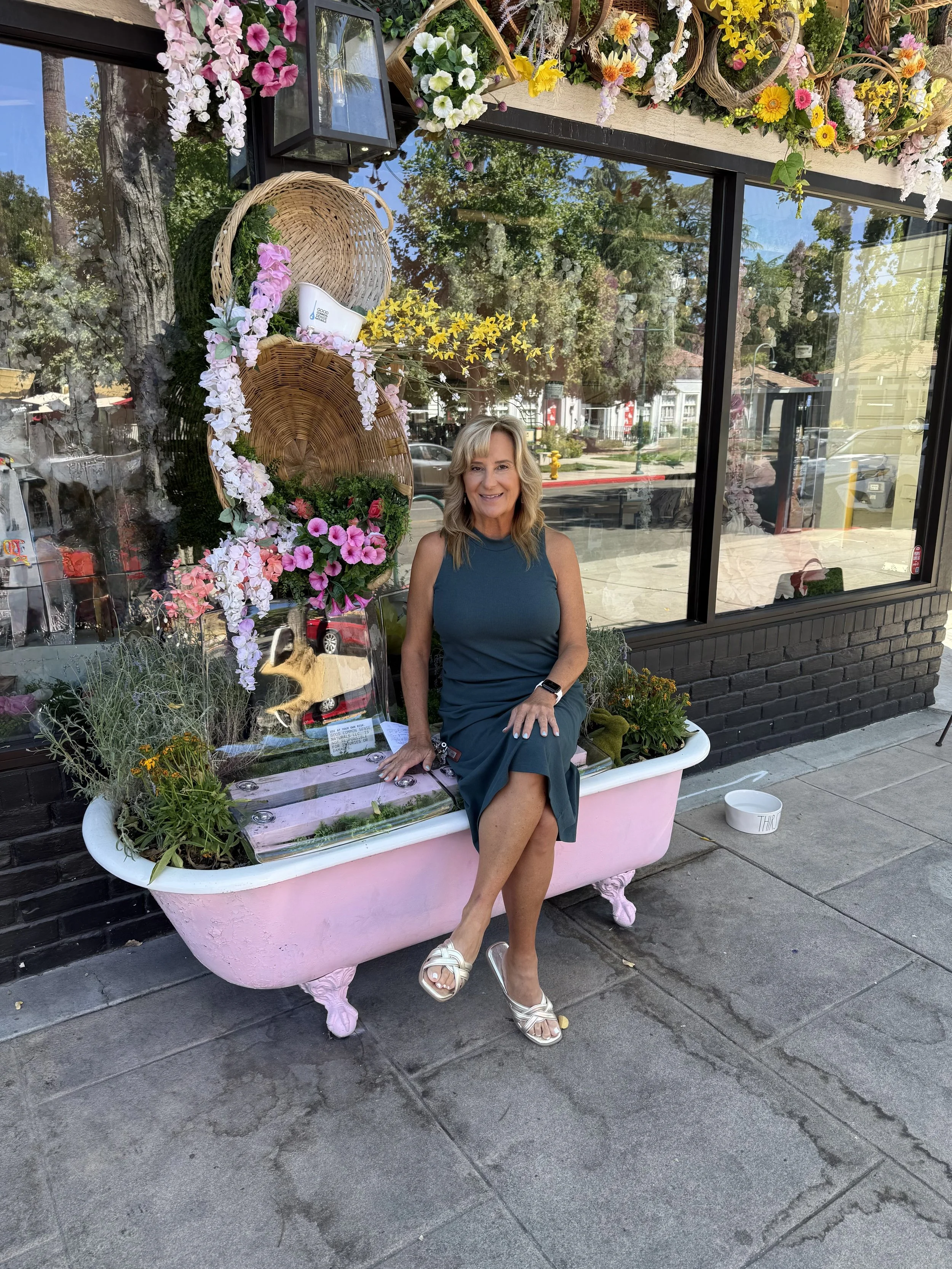 Patty Recupero posing on a vintage bathtub-style bench surrounded by fresh floral arrangements