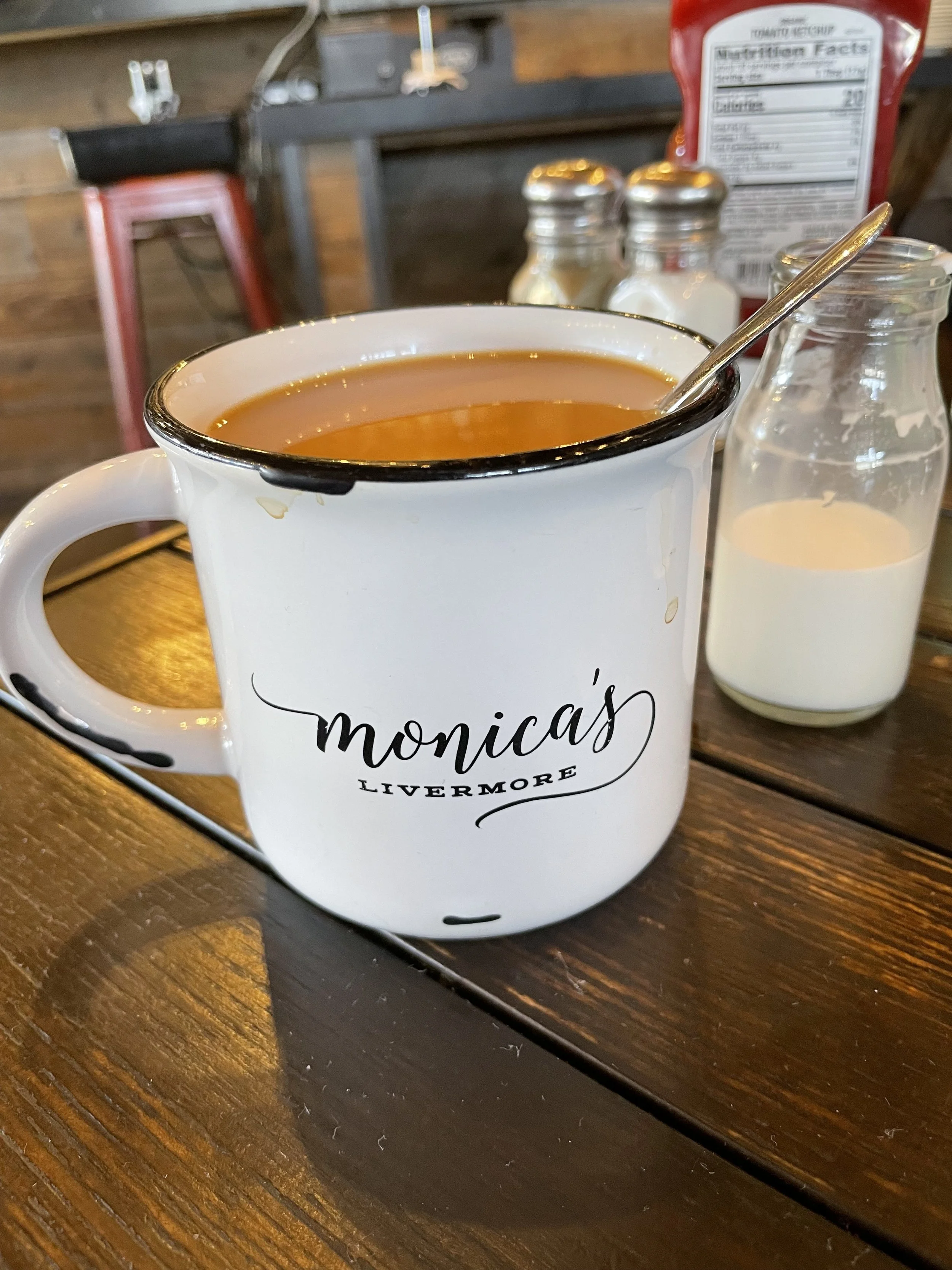 A white mug with black rim and the words 'monica's LIVERMORE' printed on it, filled with tea, placed on a wooden table. In the background, there are salt and pepper shakers, a bottle of milk, and a ketchup packet.