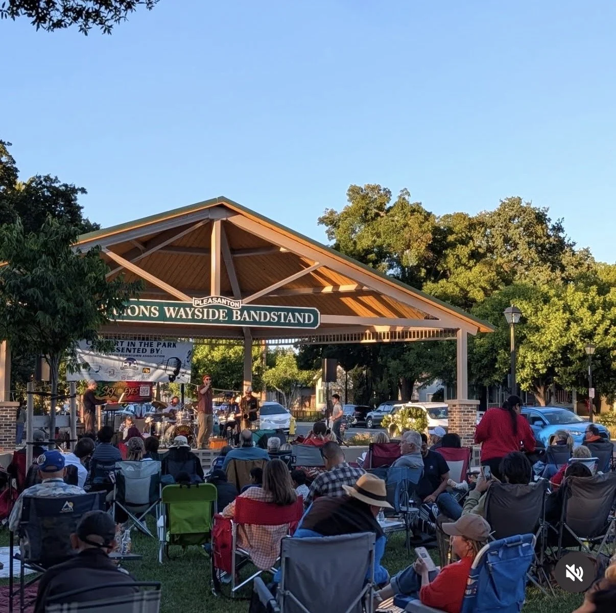 Outdoor concert at Pleasanton Lions Wayside Bandstand with a band performing on stage and audience members seated in lawn chairs during the daytime.
