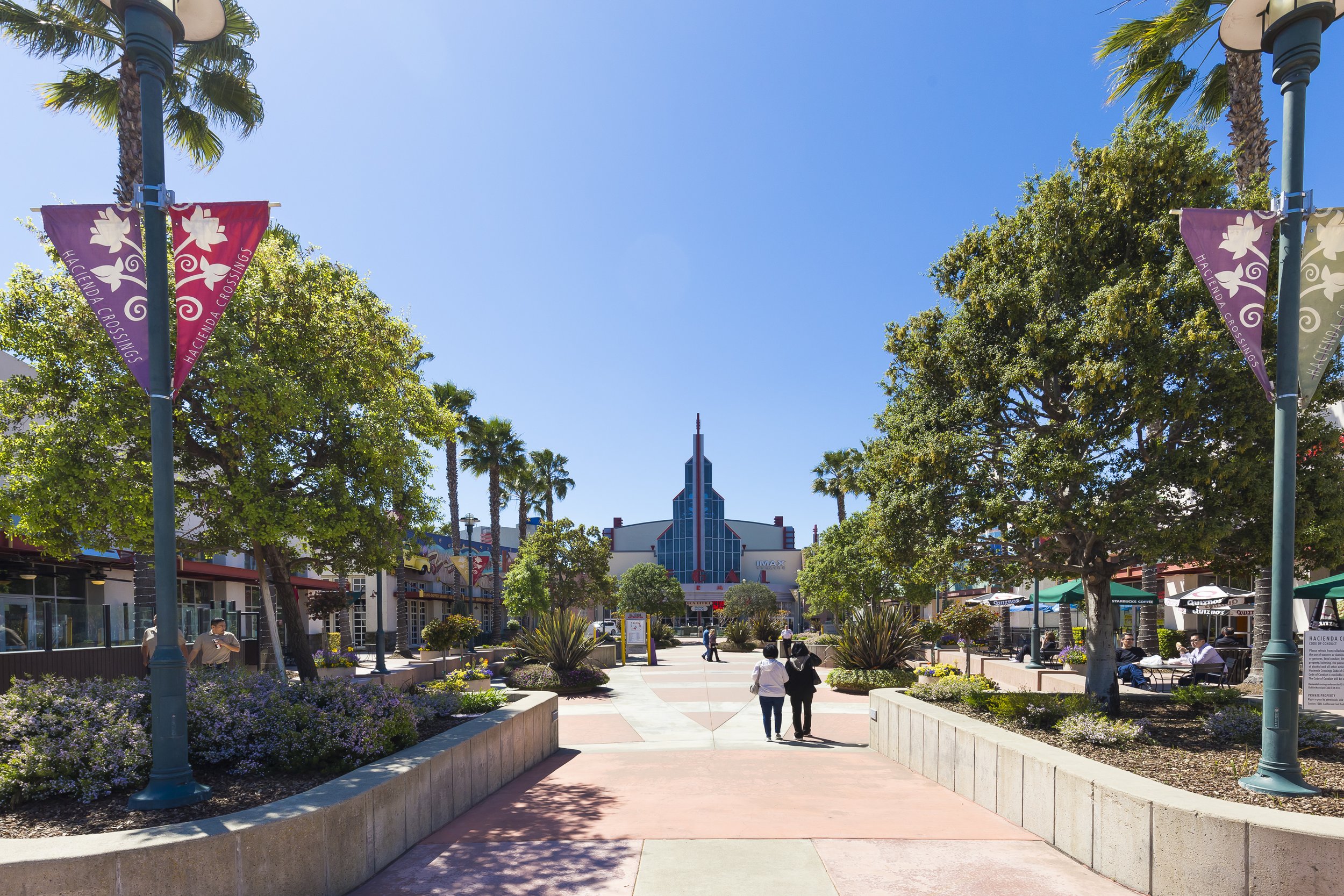 A sunny outdoor shopping mall with trees, purple flowerbeds, and various shops and cafes. Two lampposts with colorful banners are visible, and people are walking along the wide pedestrian pathway.