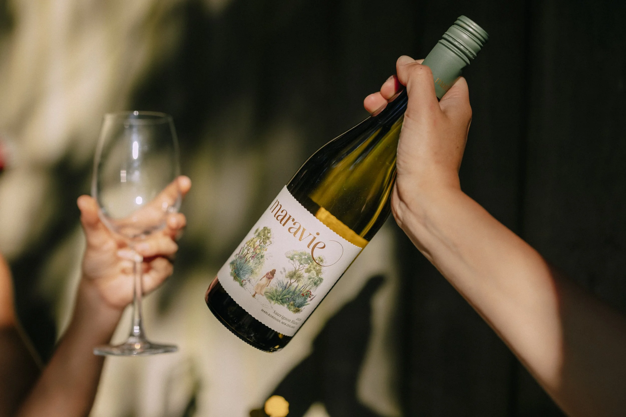 A person holding a wine bottle labeled 'Maravie' with a colorful illustration of New Zealand native trees and people, while another person holds a wine glass.