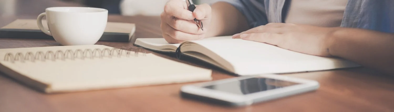 Person writing in a notebook on a wooden table with a cup, a smartphone, and other notebooks nearby.