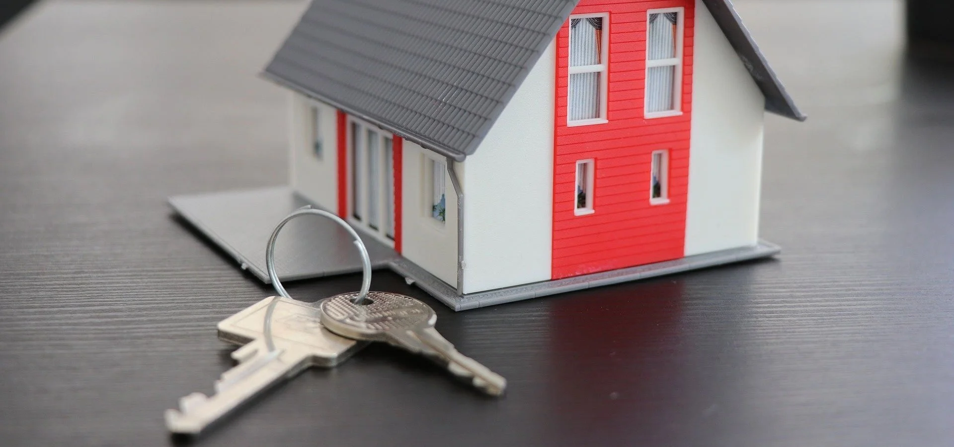 Miniature house model with red and white walls and gray roof, placed on a dark surface, with a keychain and keys in front.