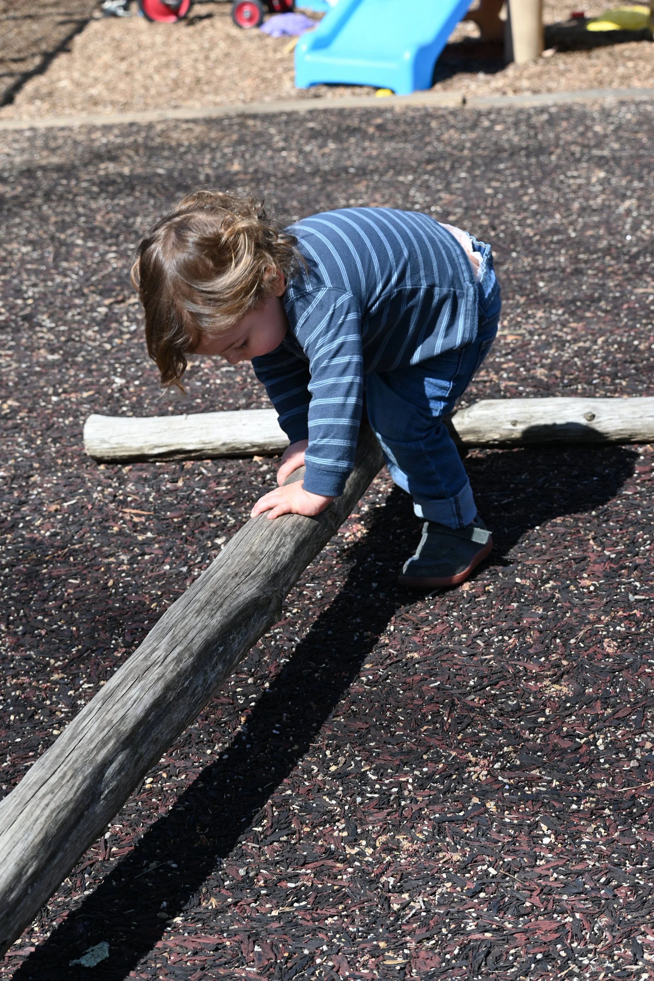 A young boy with curly brown hair wearing a blue striped long-sleeved shirt and jeans, leaning over and balancing on a weathered wooden log at a playground with a blue slide visible in the background.