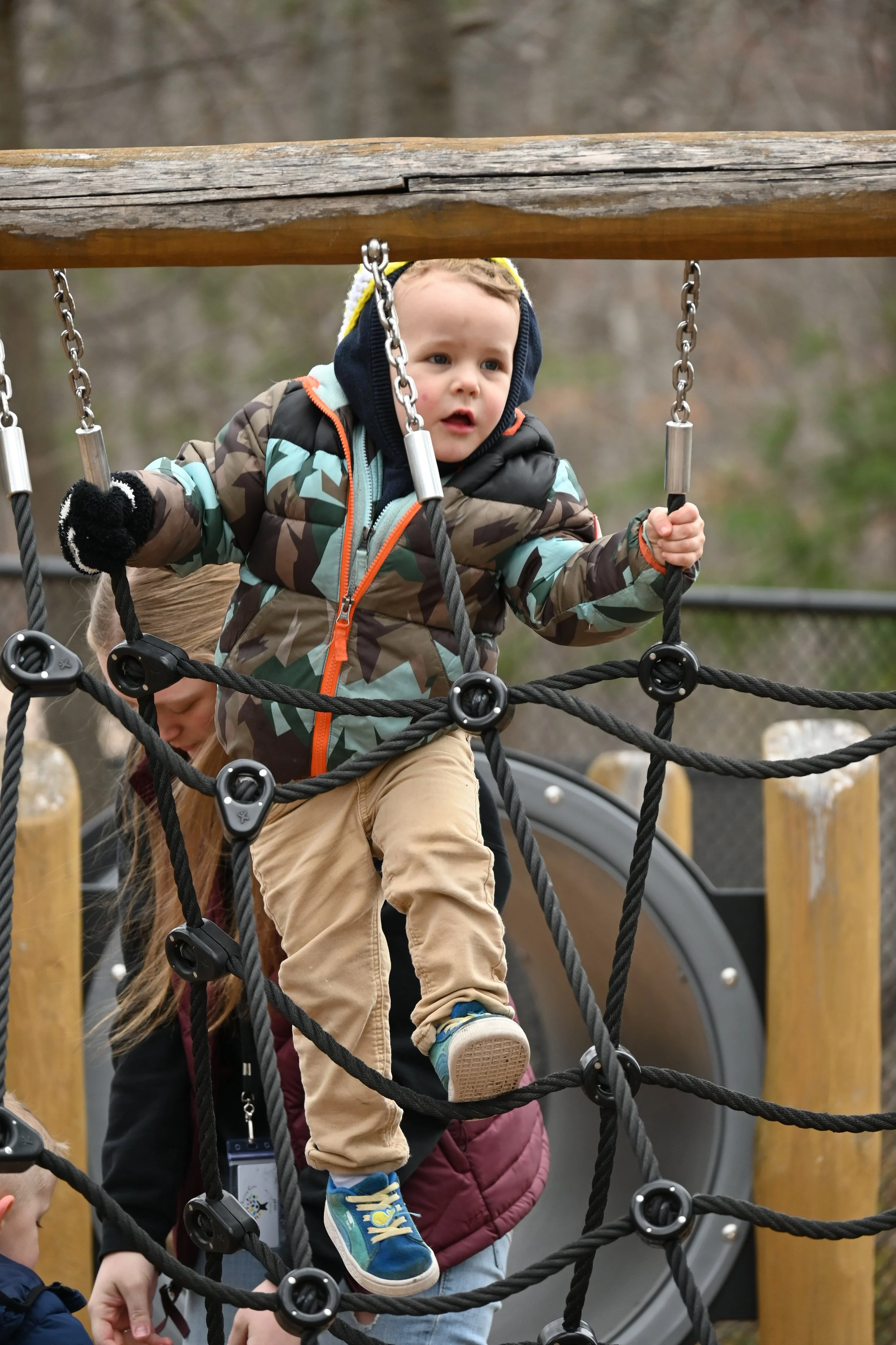 A young boy in a camouflage jacket and blue shoes climbing a net playground, with a girl in a maroon jacket assisting him.