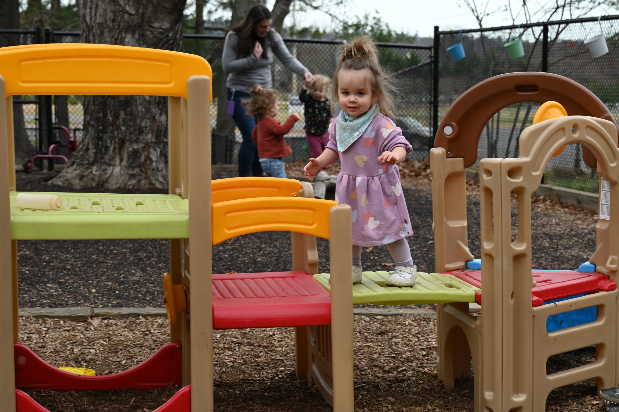 Children playing in a playground with a woman in the background. A little girl in a pink and purple dress with a heart pattern is standing on a colorful play structure, surrounded by trees and a fence.