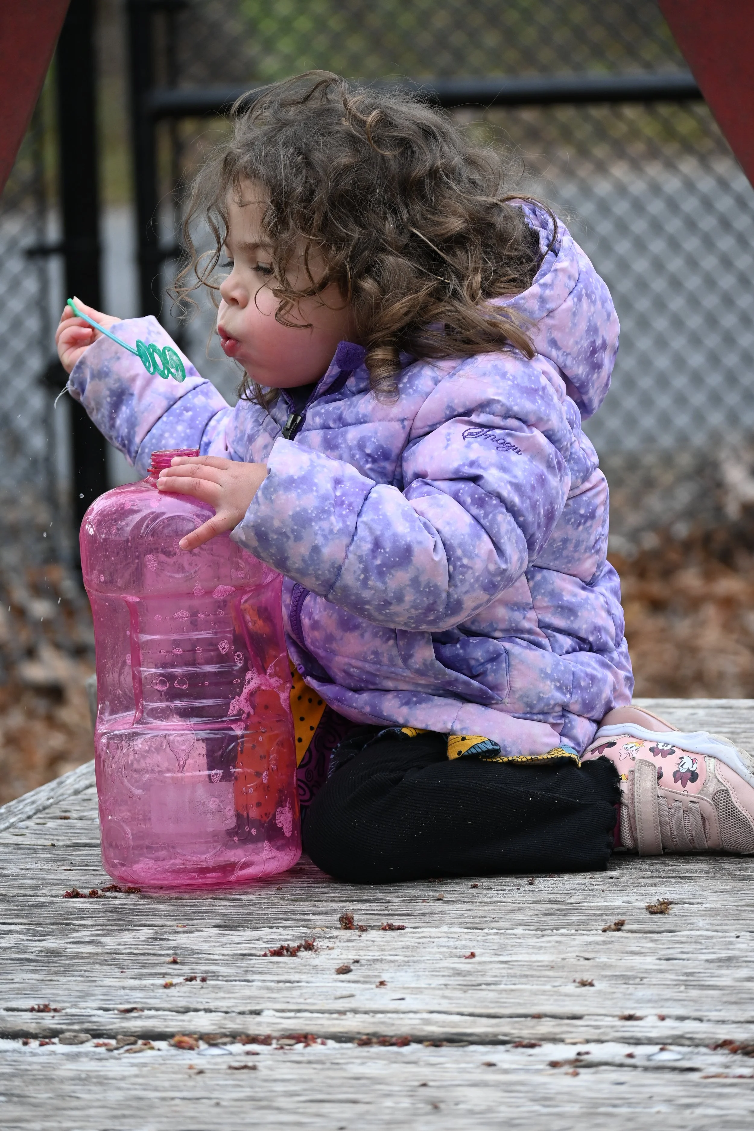 A young girl with curly brown hair sitting on a wooden picnic table outdoors, holding a pink water bottle and a bubble wand, wearing a purple tie-dye jacket, black pants, pink Minnie Mouse shoes, and patterned leggings, with a chain-link fence in the background.