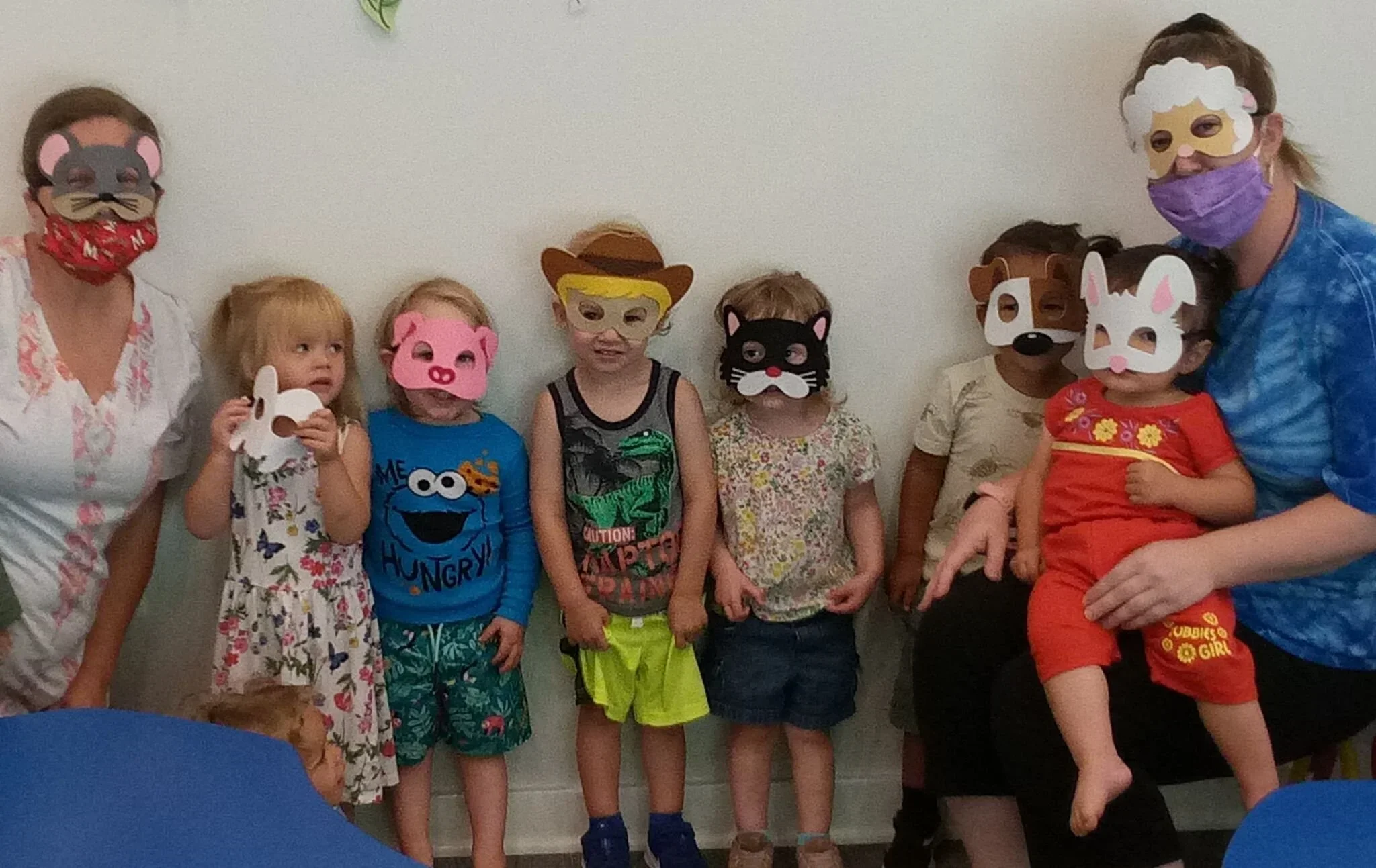 A group of children with animal masks on their faces and two adults, one on each side, in a room with a plain wall. The children are standing close together, and some are holding paper animal masks. The two adults are wearing masks—one with a bear face and the other with a sheep face—and are dressed casually.