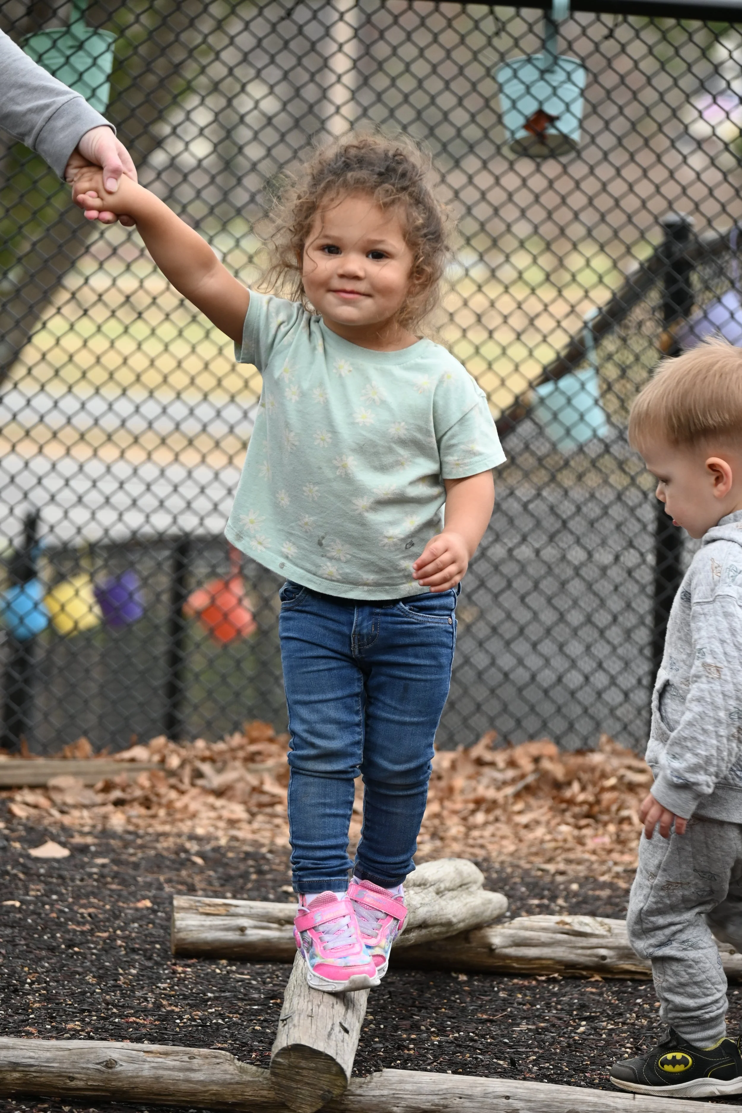 A young girl with curly hair, wearing a light green t-shirt with a daisy pattern, blue jeans, and pink sneakers, is balancing on a log in a playground area. A boy with light-colored hair, dressed in a gray hoodie with a Batman logo and gray sweatpants, stands nearby.