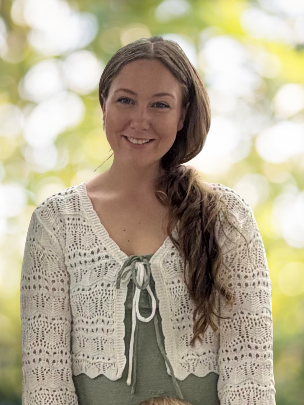 A smiling woman with long brown hair wearing a cream-colored, crochet cardigan over a green top, standing outdoors with blurred green and yellow foliage in the background.