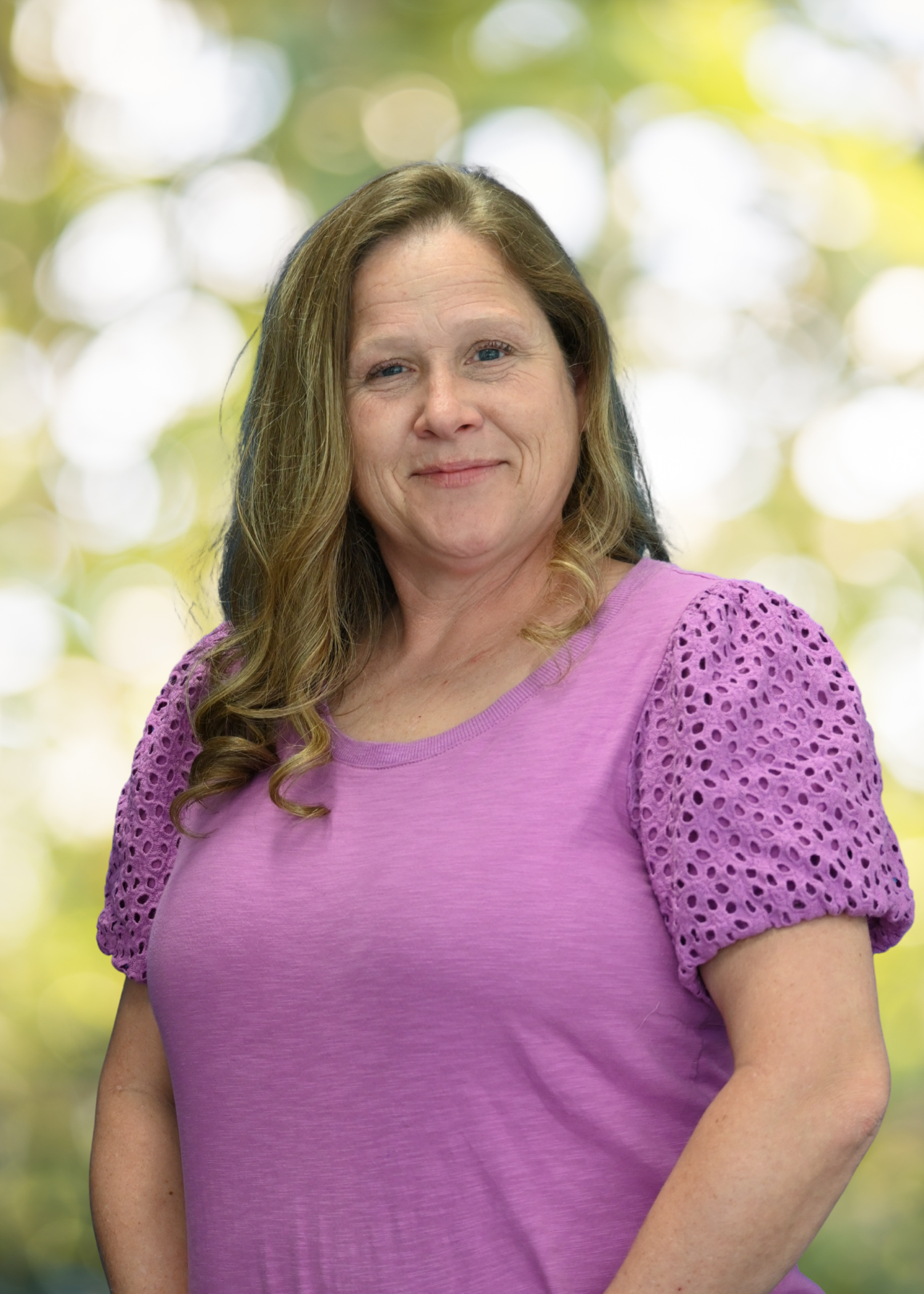 A woman with long brown hair, wearing glasses and a gray t-shirt, smiling, standing outdoors with blurred green and yellow bokeh background.