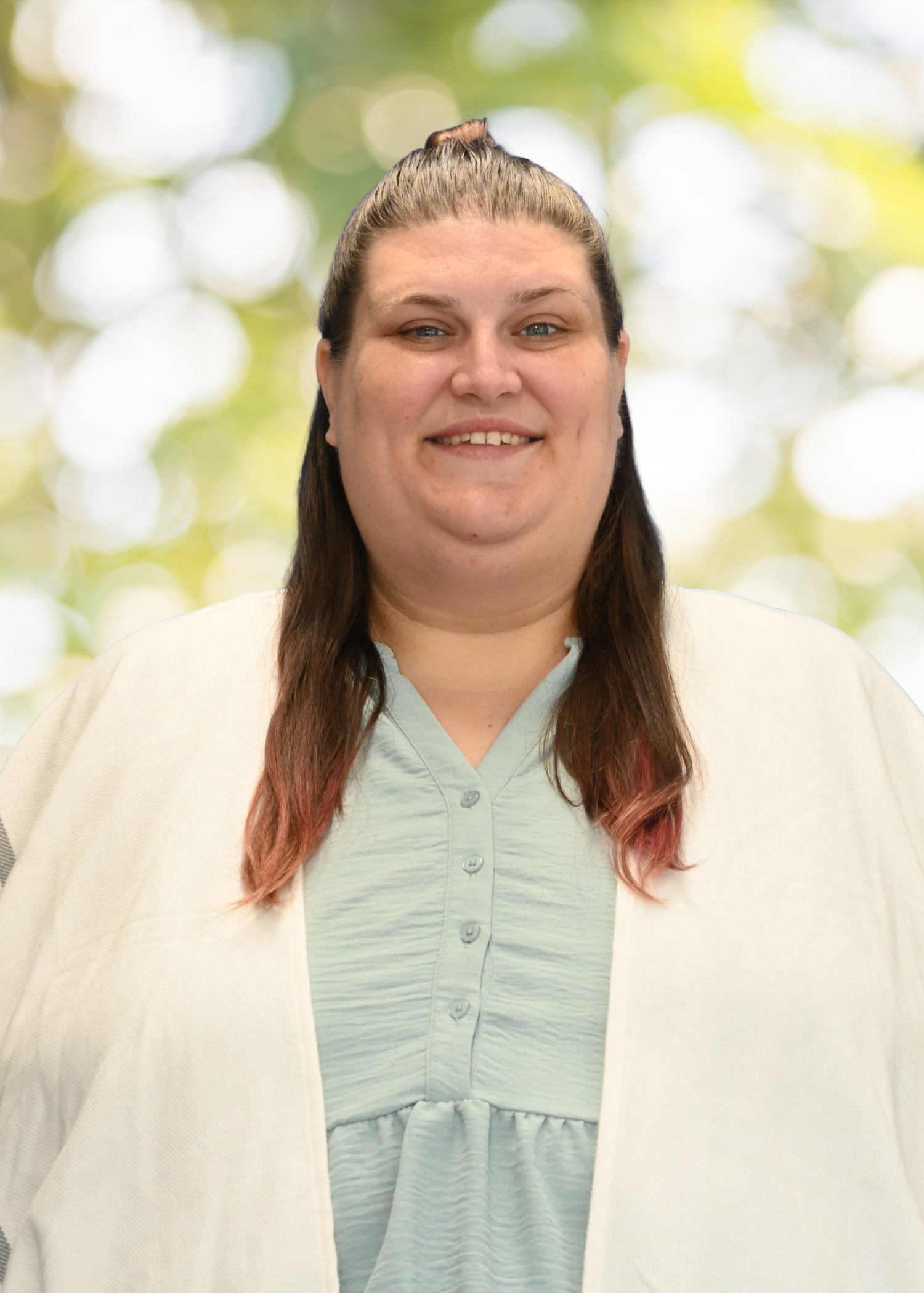 A woman with brown hair, streaked with pink at the ends, smiling, wearing a light green hospital gown and a cream-colored cardigan, standing outdoors with blurred green and yellow foliage in the background.