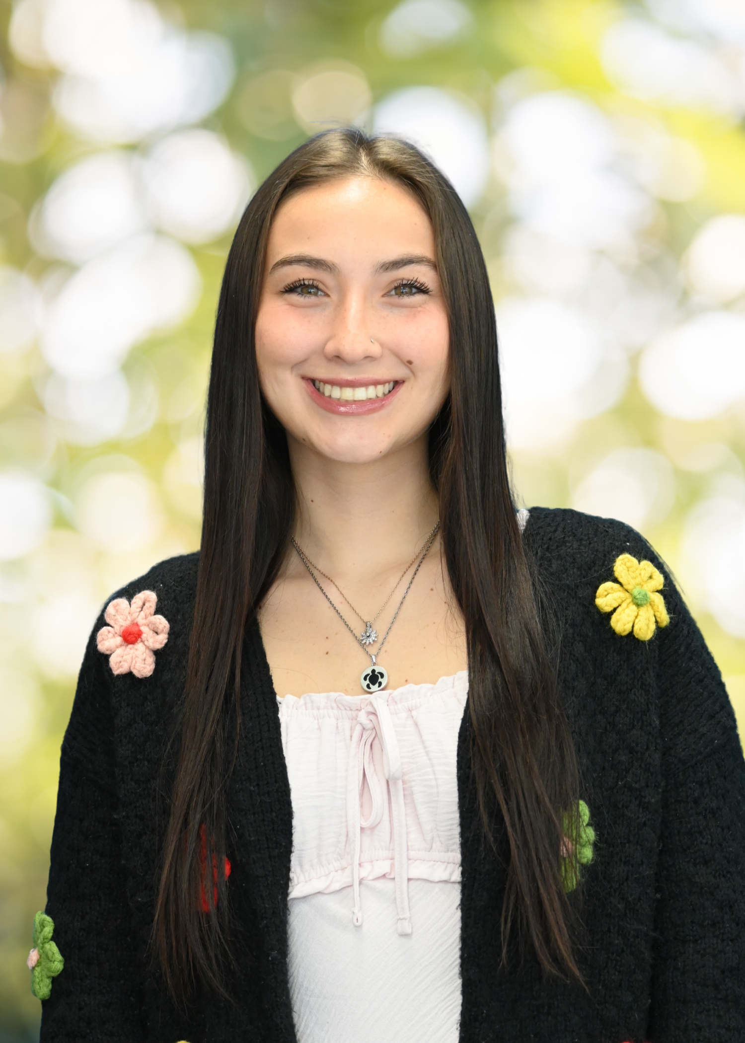 A young woman with long red hair, blue eyes, and a bright smile, wearing a white top, burgundy cardigan, and a silver necklace with a dog tag pendant, standing outdoors with bokeh trees and sunlight in the background.