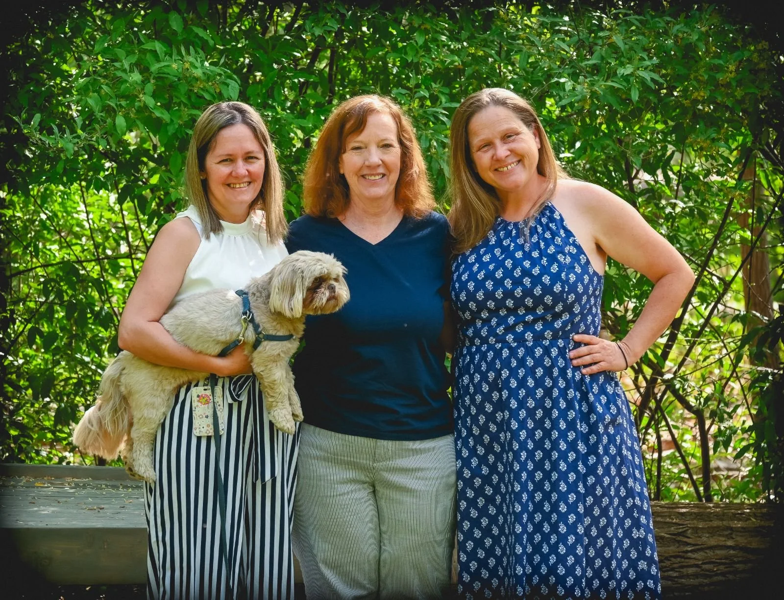 Three women smiling outdoors, with green foliage in the background. The woman on the left holds a small, light-colored dog in her arm. The woman in the middle has curly red hair, and the woman on the right is wearing a blue patterned dress, with her hand on her hip.