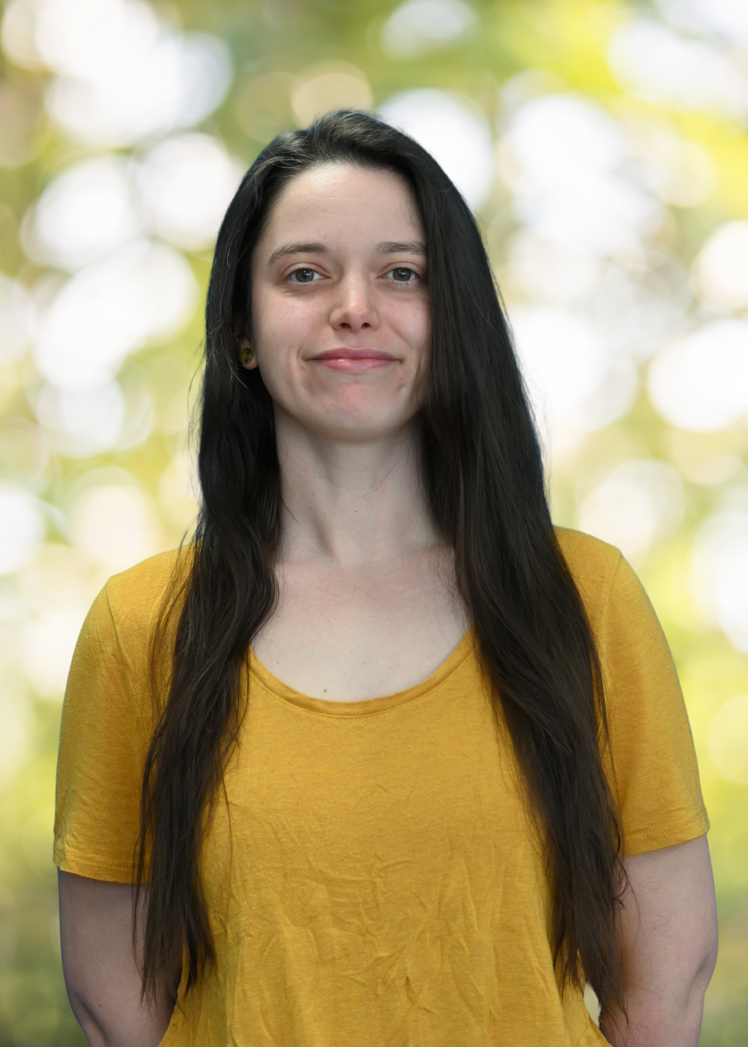 A woman with dark brown hair wearing a beige sweater over a white collared shirt, standing outdoors with a blurred background of trees and yellow-green light.
