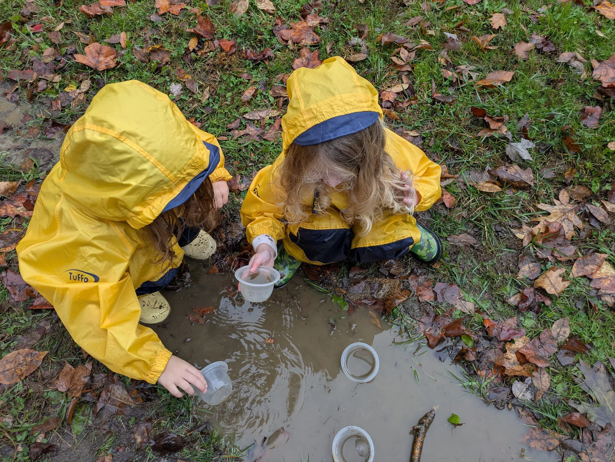 Two children in yellow waterproof jackets are crouched down, playing in a muddy puddle on a grassy area with fallen autumn leaves. They are holding plastic cups, possibly engaged in a water play activity.