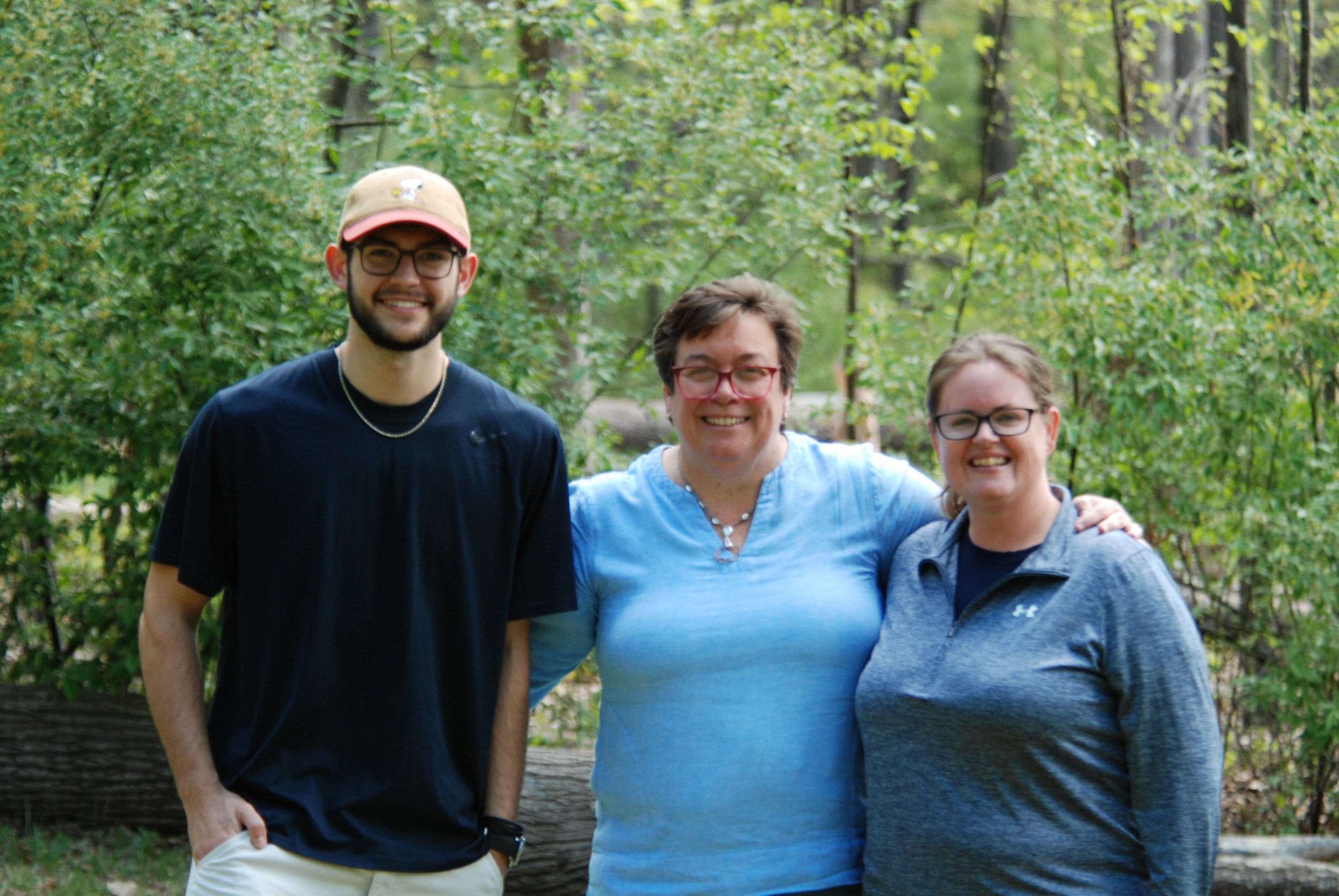 Three smiling people standing in a wooded park, with two women and one man. The man is wearing glasses, a tan cap, and a black shirt. The woman in the middle has short gray hair, glasses, and a light blue shirt. The woman on the right has glasses, short gray hair, and a dark blue jacket. They are standing close together with a background of green trees and foliage.