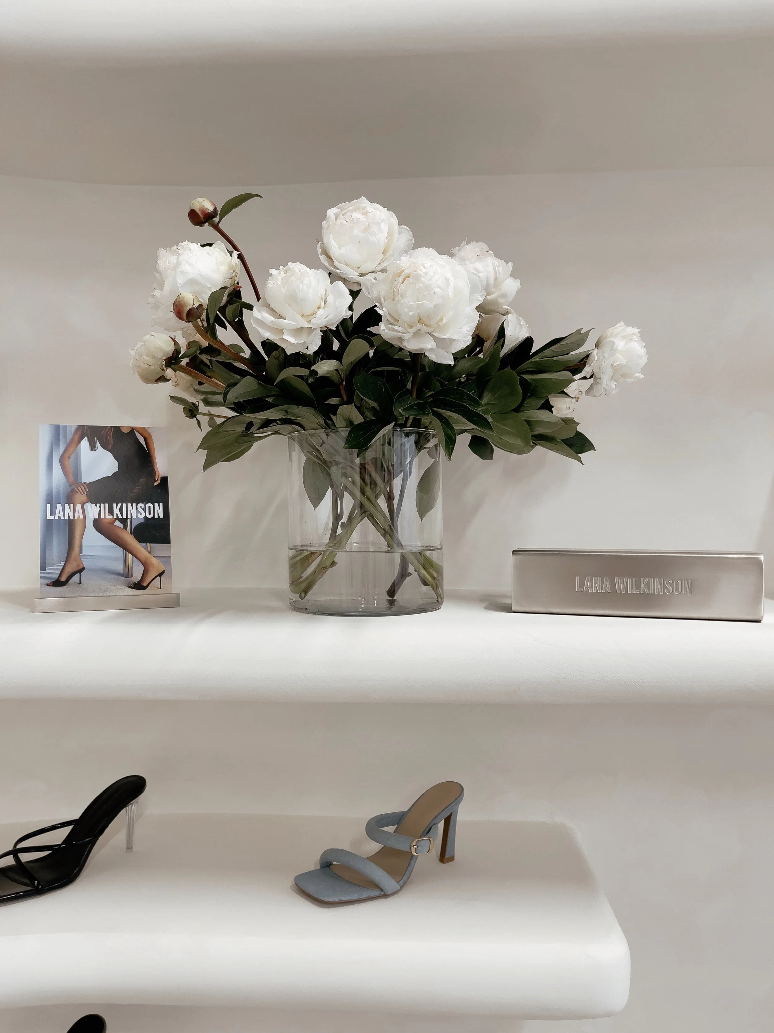 A display of a pair of light blue heels and black stilettos, along with a sign featuring the name Lana Wilkinson, and a glass vase with white peony flowers on a white shelf.