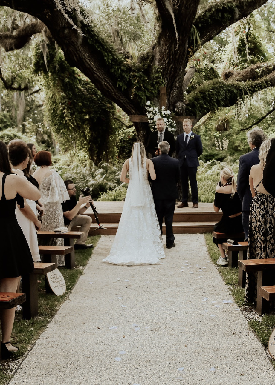 A wedding ceremony taking place outdoors in front of a large tree with green foliage. The bride in a white lace wedding dress and veil is walking down the aisle with an older man, likely her father. The groom stands nearby with a groomsman, all dressed in dark suits. Guests are seated on wooden benches on either side of the aisle, watching the ceremony amidst lush greenery.