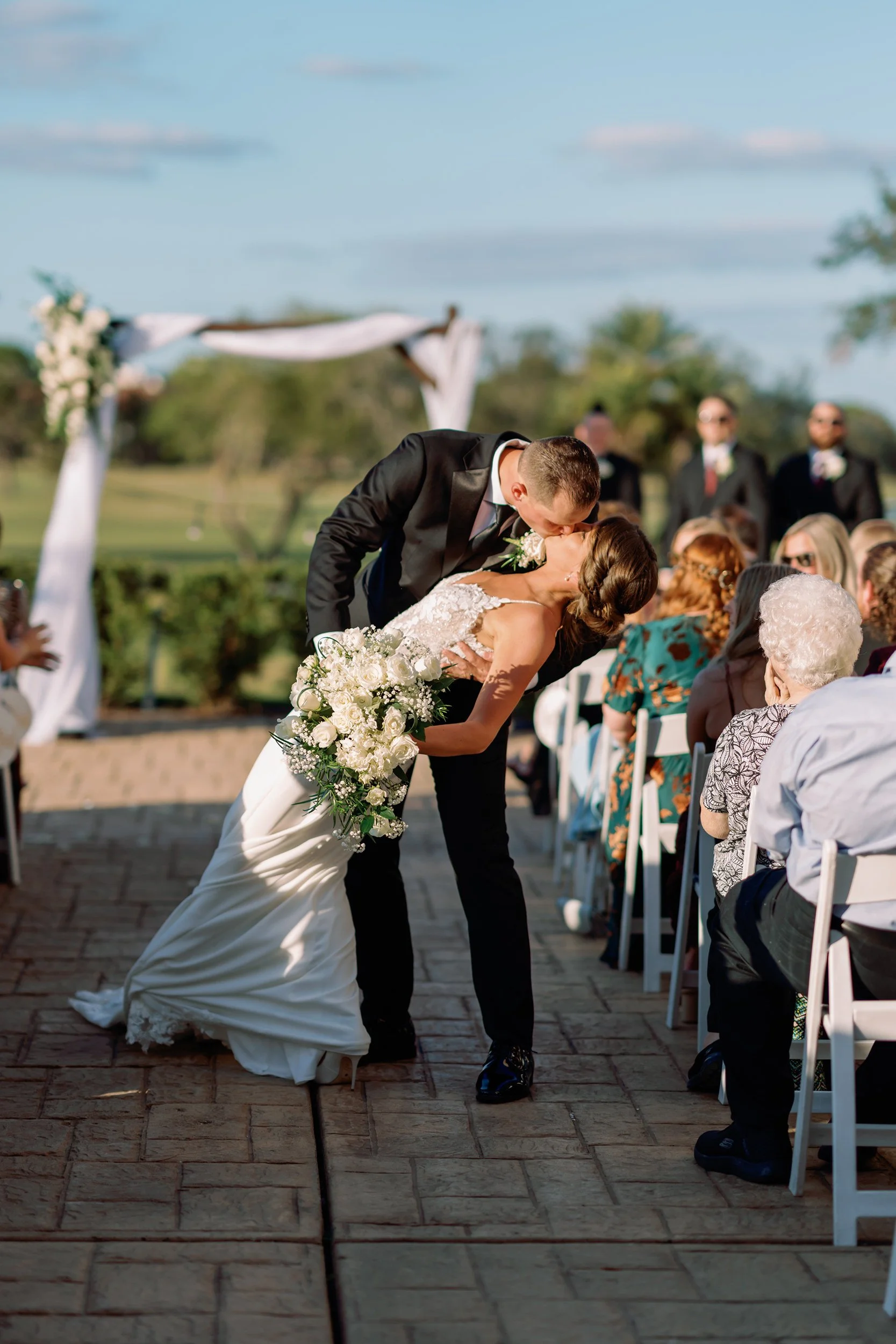 A bride and groom sharing a kiss during their outdoor wedding ceremony, with guests seated on white chairs and a decorated arch in the background on a sunny day.