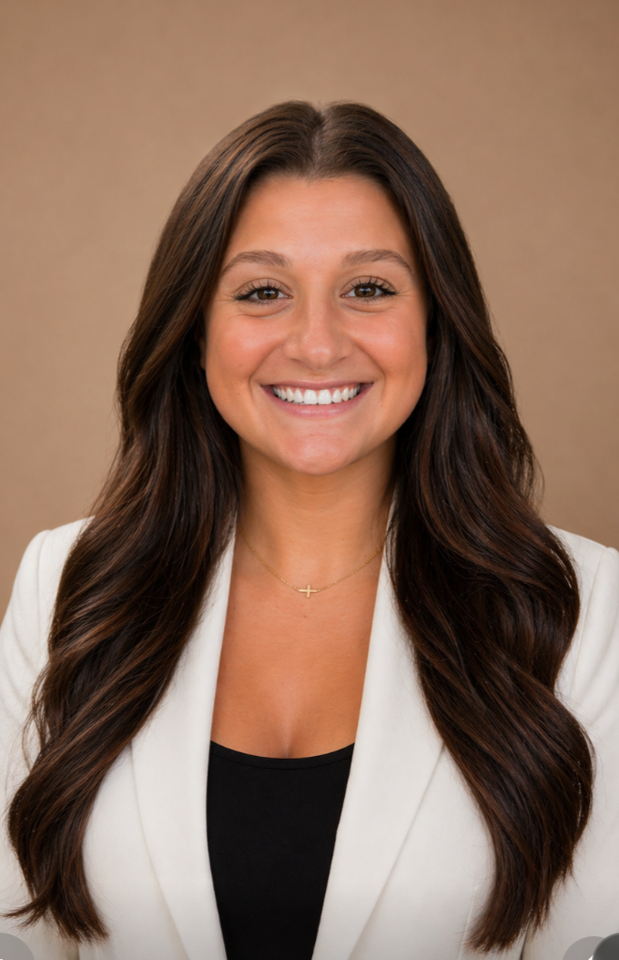 A woman with long brown hair, smiling, wearing a white blazer and a black top, with a gold Cross necklace, against a neutral background.