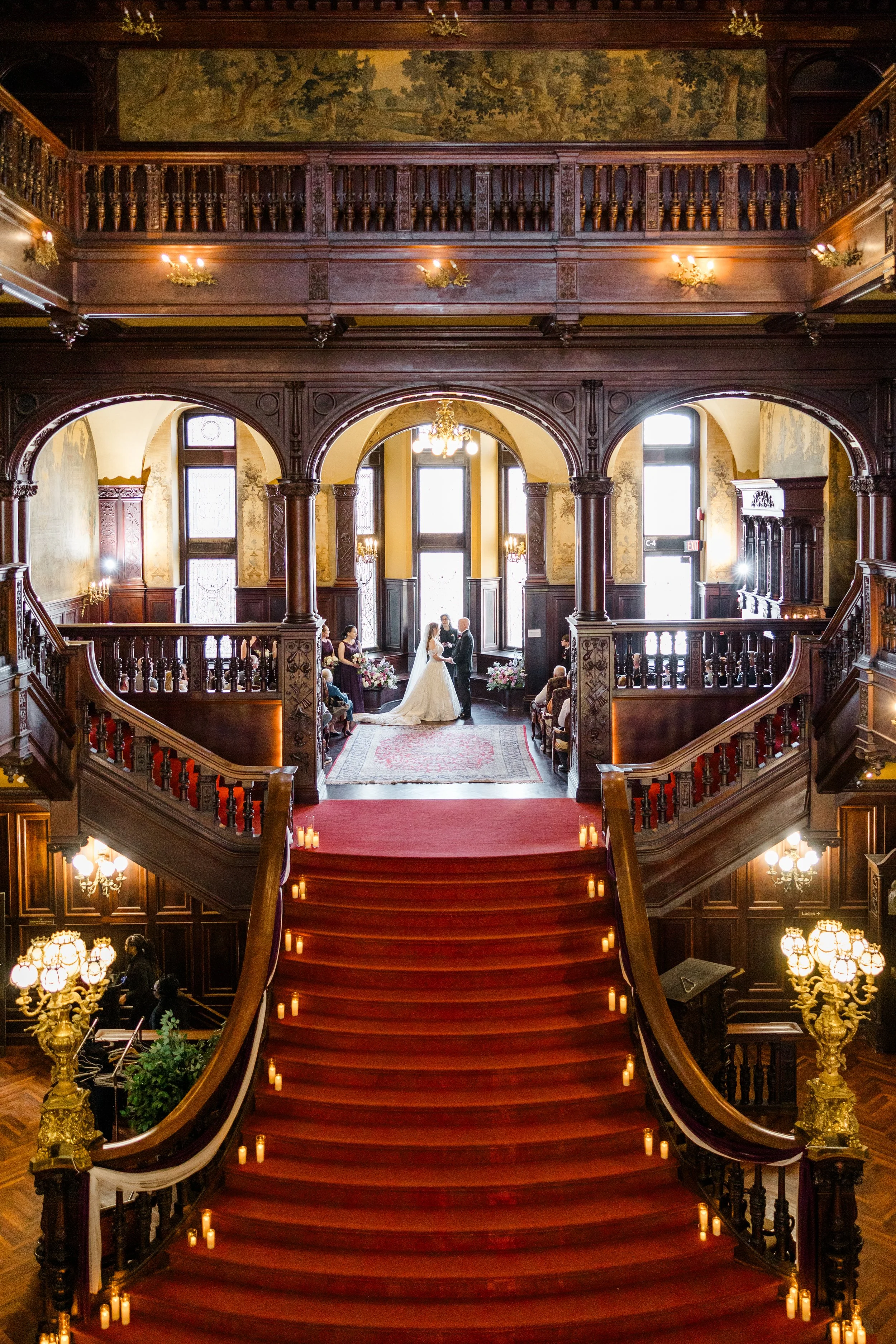 A grand, elegantly decorated wedding venue with a large staircase leading to a second floor. In the background, a bride and groom are exchanging vows under large windows, surrounded by floral arrangements. The area is illuminated by chandeliers and candlelight, with wood-paneled walls and ornate details.